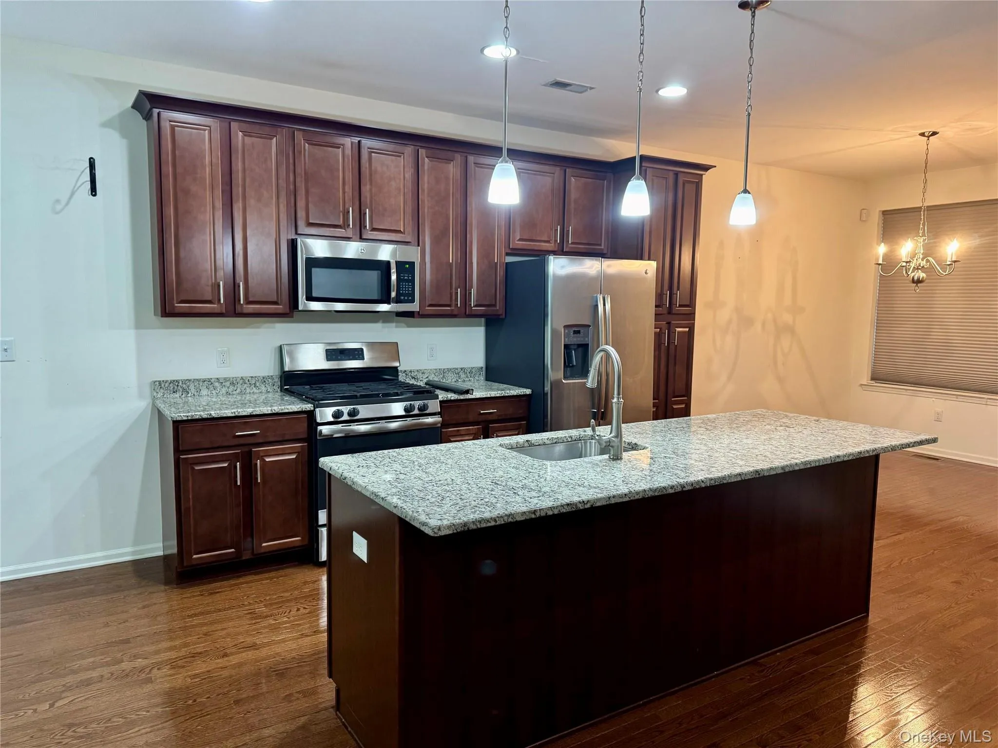 Kitchen featuring appliances with stainless steel finishes, dark wood-style floors, hanging light fixtures, light stone countertops, and a kitchen island with sink Kitchen featuring appliances with stainless steel finishes, dark wood-style floors, hanging light fixtures, light stone countertops, and a kitchen island with sink