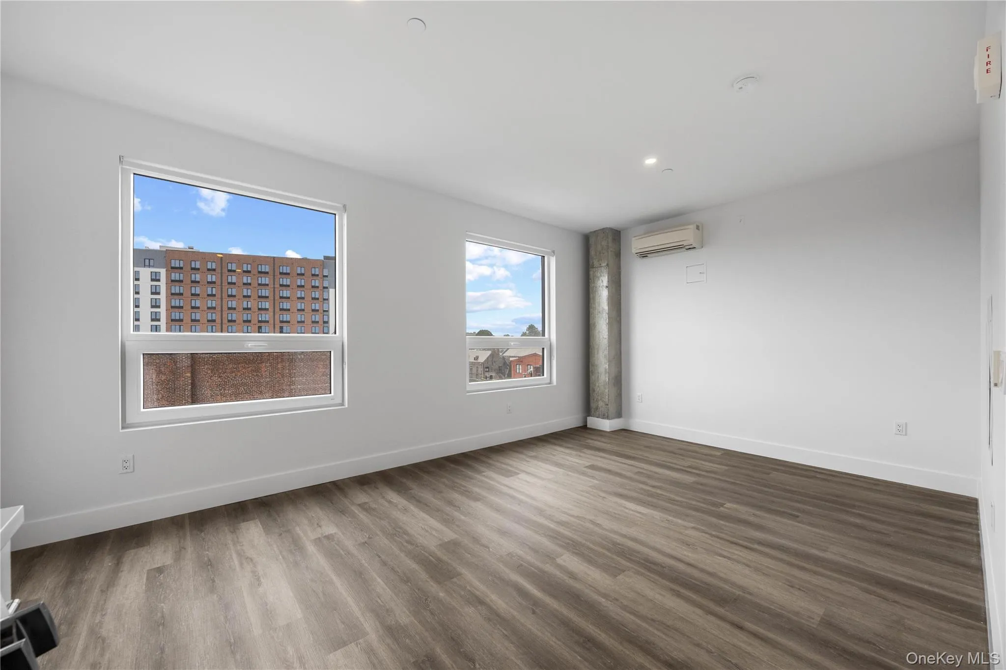 Spacious room featuring wood-finish flooring, two large windows, white walls, and a visible concrete support column Spacious room featuring wood-finish flooring, two large windows, white walls, and a visible concrete support column