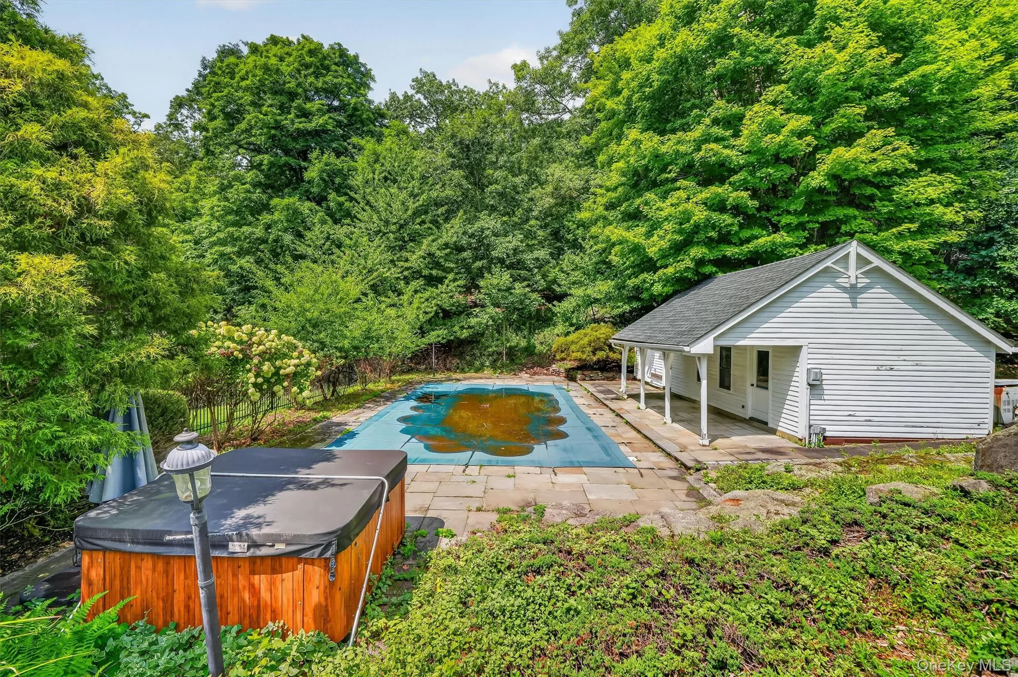 Outdoor swimming pool with surrounding stone patio, white siding pool house with covered porch, and wood-sided hot tub Outdoor swimming pool with surrounding stone patio, white siding pool house with covered porch, and wood-sided hot tub