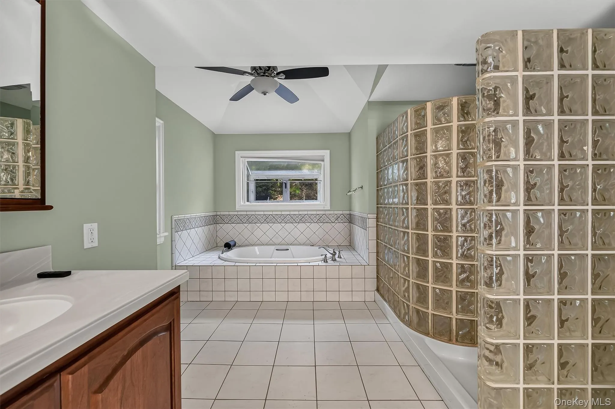Recessed tub with tile surround, white tile flooring, ceiling fan, and curved glass block shower wall, Jetted Shower behind it. Recessed tub with tile surround, white tile flooring, ceiling fan, and curved glass block shower wall, Jetted Shower behind it.