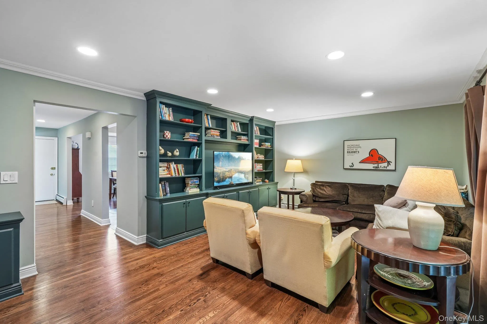 Living area featuring wood-finish flooring, built-in shelving with integrated media space, recessed lighting, crown molding, and an arched doorway Living area featuring wood-finish flooring, built-in shelving with integrated media space, recessed lighting, crown molding, and an arched doorway