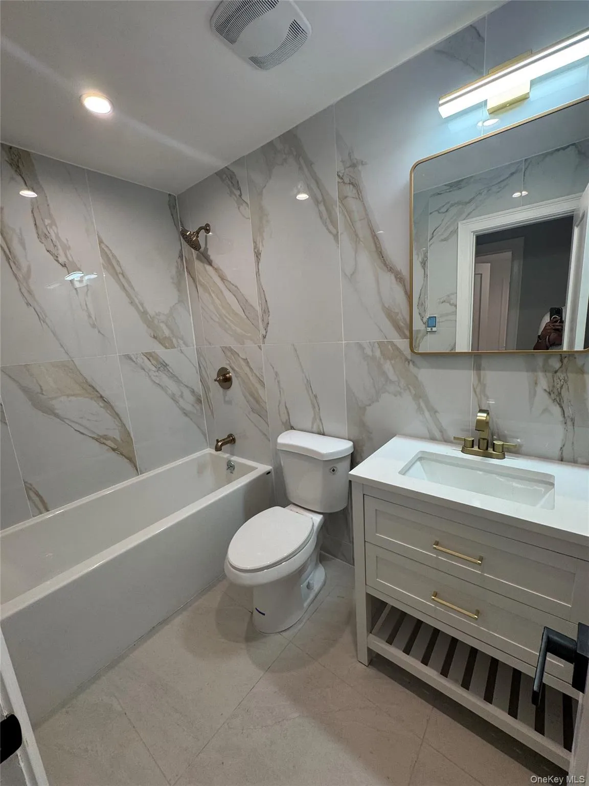 Bathroom featuring large-format wall tiling with subtle veining, a built-in tub with a shower fixture, a contemporary vanity with a white countertop and gold-toned hardware, a rectangular mirror with a gold-toned frame, and recessed ceiling lighting Bathroom featuring large-format wall tiling with subtle veining, a built-in tub with a shower fixture, a contemporary vanity with a white countertop and gold-toned hardware, a rectangular mirror with a gold-toned frame, and recessed ceiling lighting