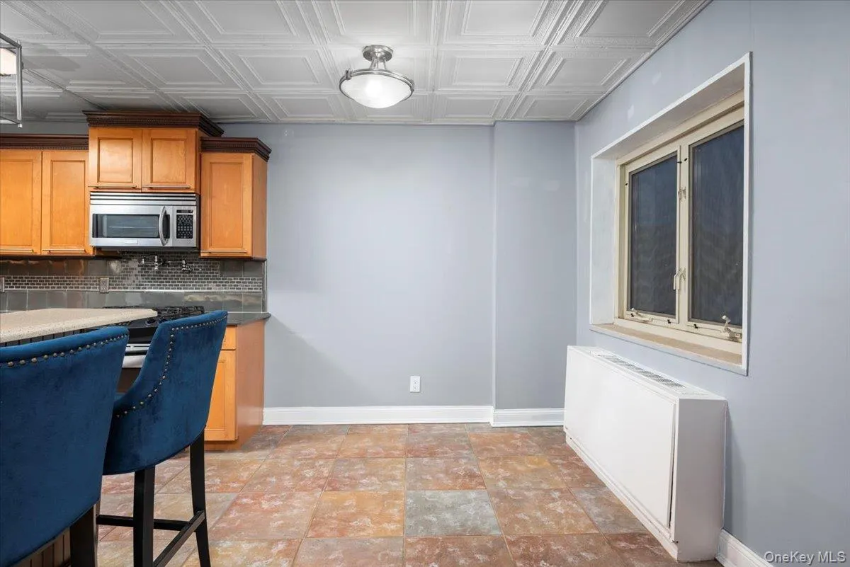 Kitchen area featuring a patterned ceiling, wood cabinetry, a built-in microwave, tile backsplash, and a window with a radiator below Kitchen area featuring a patterned ceiling, wood cabinetry, a built-in microwave, tile backsplash, and a window with a radiator below