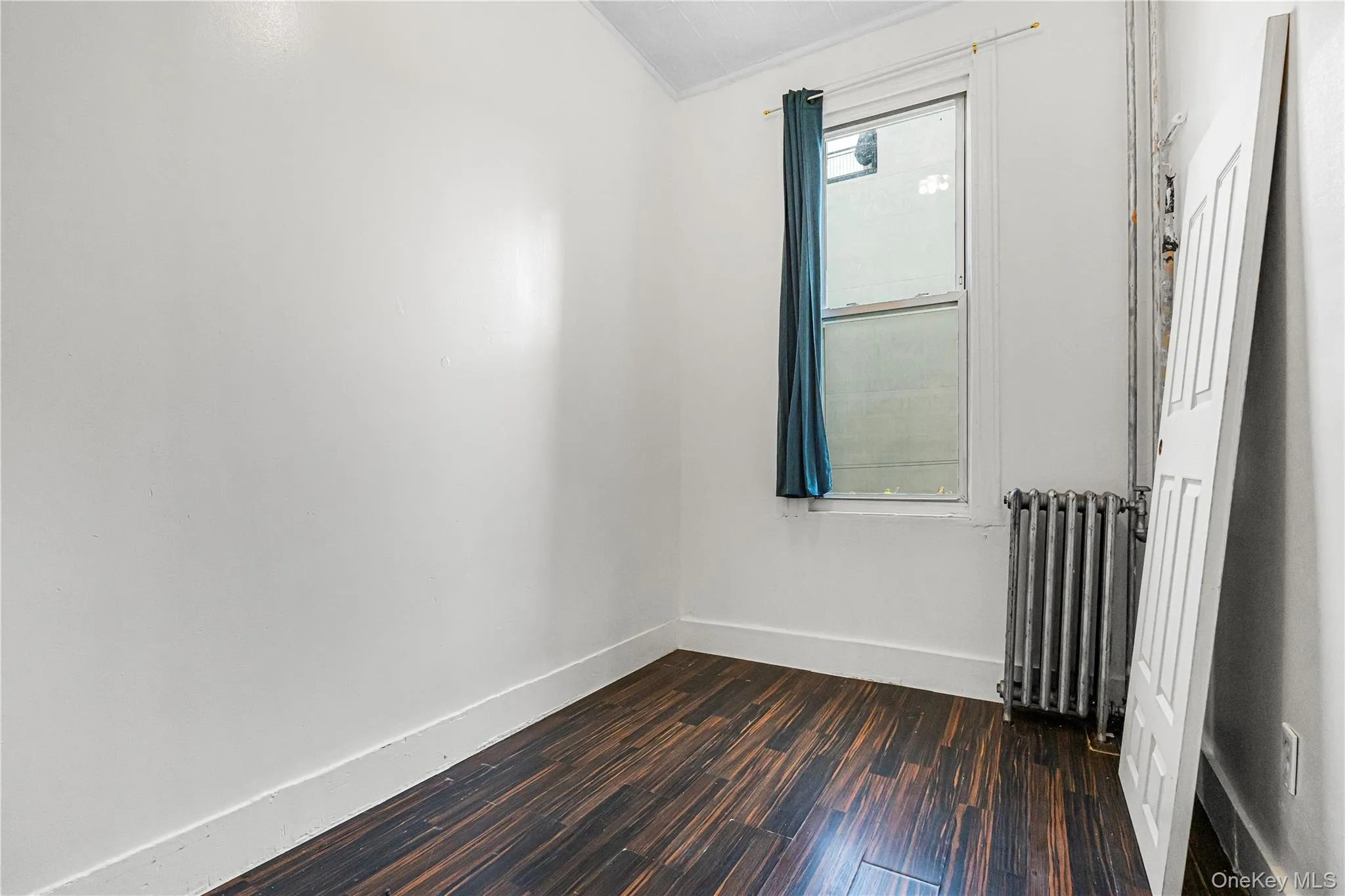 Bright room featuring wood-finish flooring, white walls, and a window with a dark curtain Bright room featuring wood-finish flooring, white walls, and a window with a dark curtain
