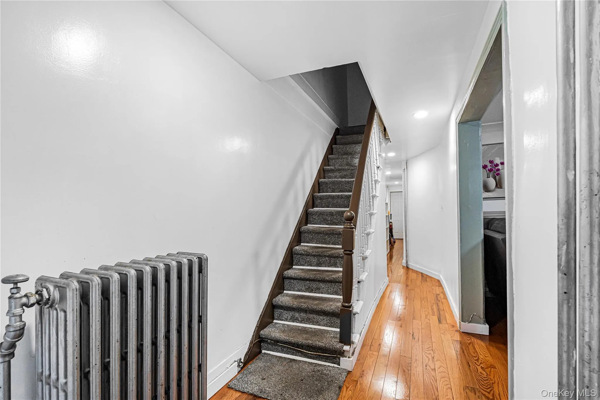 Hallway featuring hardwood flooring, a carpeted staircase with a white spindle banister, recessed lighting, and a cast iron radiator Hallway featuring hardwood flooring, a carpeted staircase with a white spindle banister, recessed lighting, and a cast iron radiator
