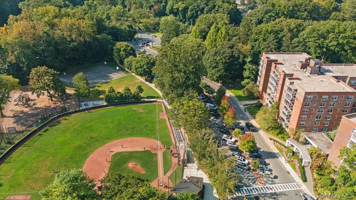 Expansive baseball field with manicured grass and dirt infield, bordered by mature trees and residential buildings Expansive baseball field with manicured grass and dirt infield, bordered by mature trees and residential buildings