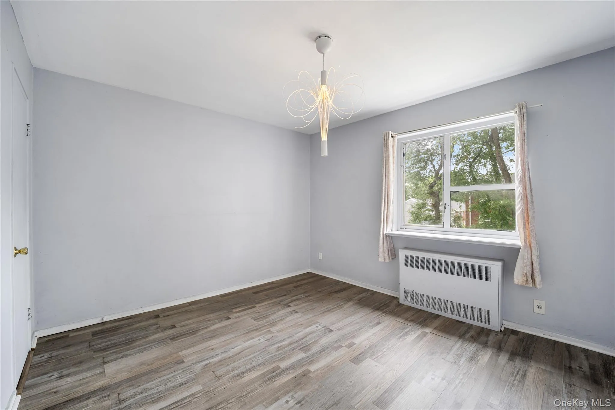 Room featuring light gray walls, wood-finish flooring, a window with curtains, a built-in radiator, and a decorative ceiling light fixture Room featuring light gray walls, wood-finish flooring, a window with curtains, a built-in radiator, and a decorative ceiling light fixture