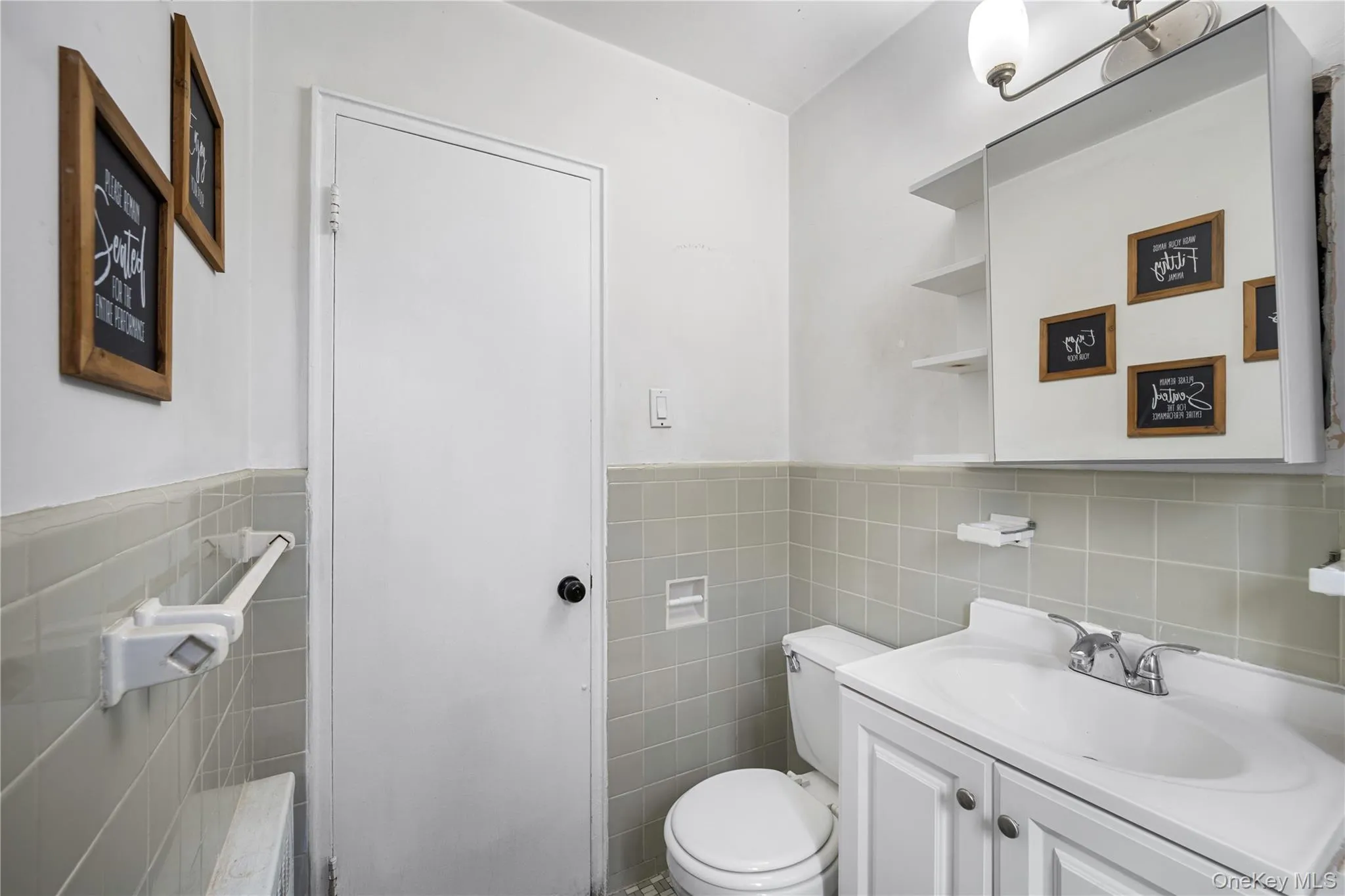 Bathroom featuring a white vanity with a single basin sink, light gray wall tiling, a mirrored medicine cabinet with open shelving, and a wall-mounted light fixture Bathroom featuring a white vanity with a single basin sink, light gray wall tiling, a mirrored medicine cabinet with open shelving, and a wall-mounted light fixture