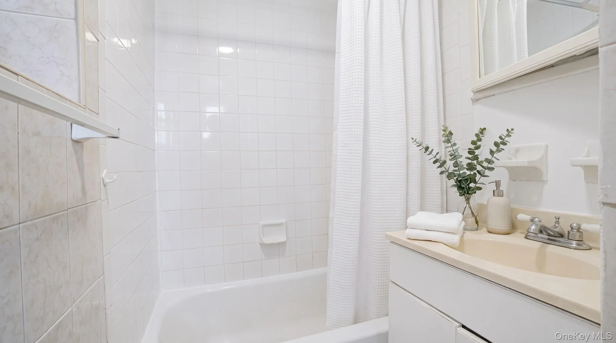 Full bathroom featuring a white bathtub and shower with white square wall tiling, a vanity with an integrated sink, and a chrome two-handle faucet Full bathroom featuring a white bathtub and shower with white square wall tiling, a vanity with an integrated sink, and a chrome two-handle faucet