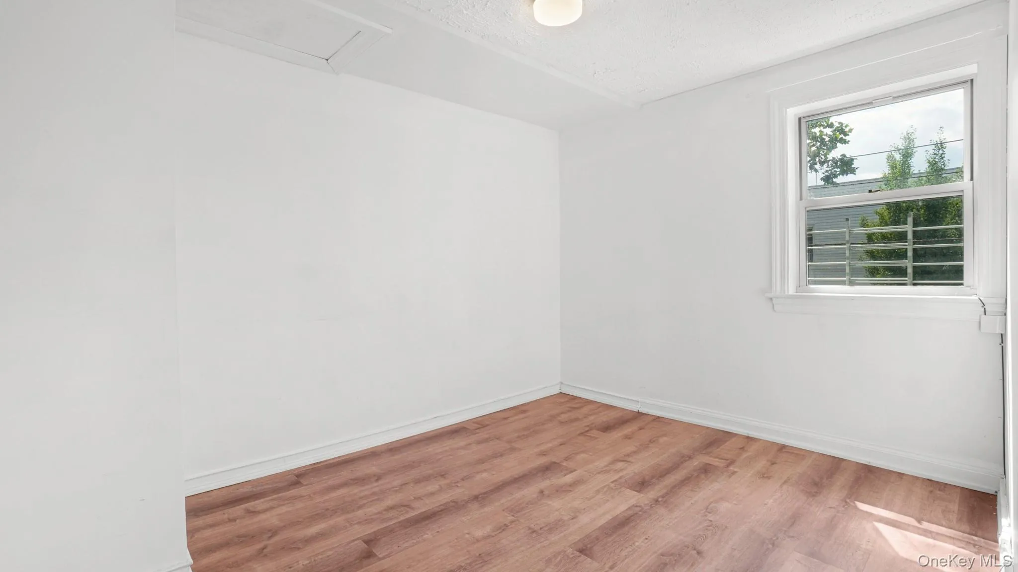 Bright room featuring wood-finish flooring, white baseboards, a single window with security bars, and a flush-mount ceiling light fixture Bright room featuring wood-finish flooring, white baseboards, a single window with security bars, and a flush-mount ceiling light fixture