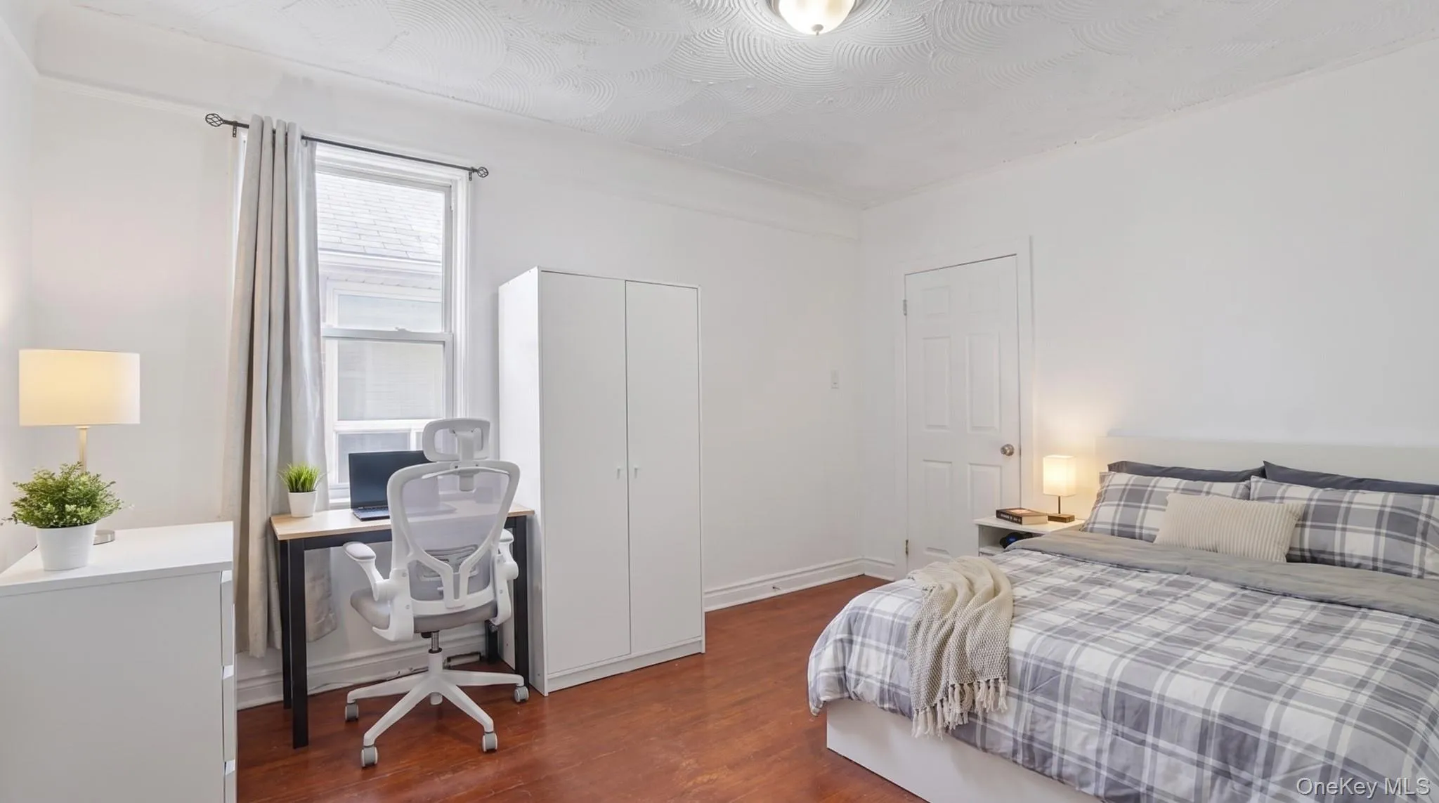Bright room featuring wood-finish flooring, a window with curtains, and overhead lighting Bright room featuring wood-finish flooring, a window with curtains, and overhead lighting