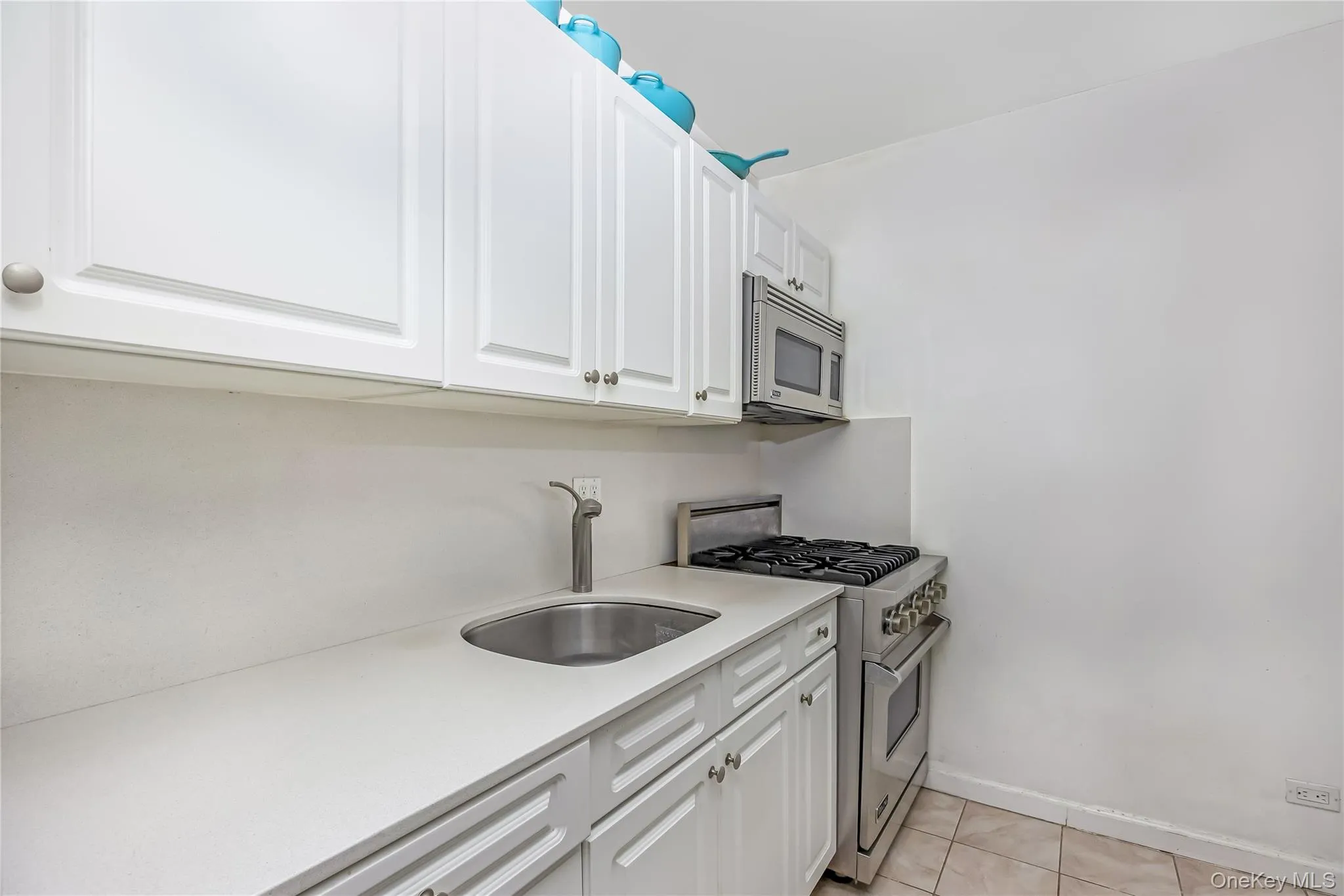 White shaker-style cabinetry with silver hardware, light-toned countertop, undermount stainless steel sink with gooseneck faucet, stainless steel microwave, and stainless steel range with oven White shaker-style cabinetry with silver hardware, light-toned countertop, undermount stainless steel sink with gooseneck faucet, stainless steel microwave, and stainless steel range with oven