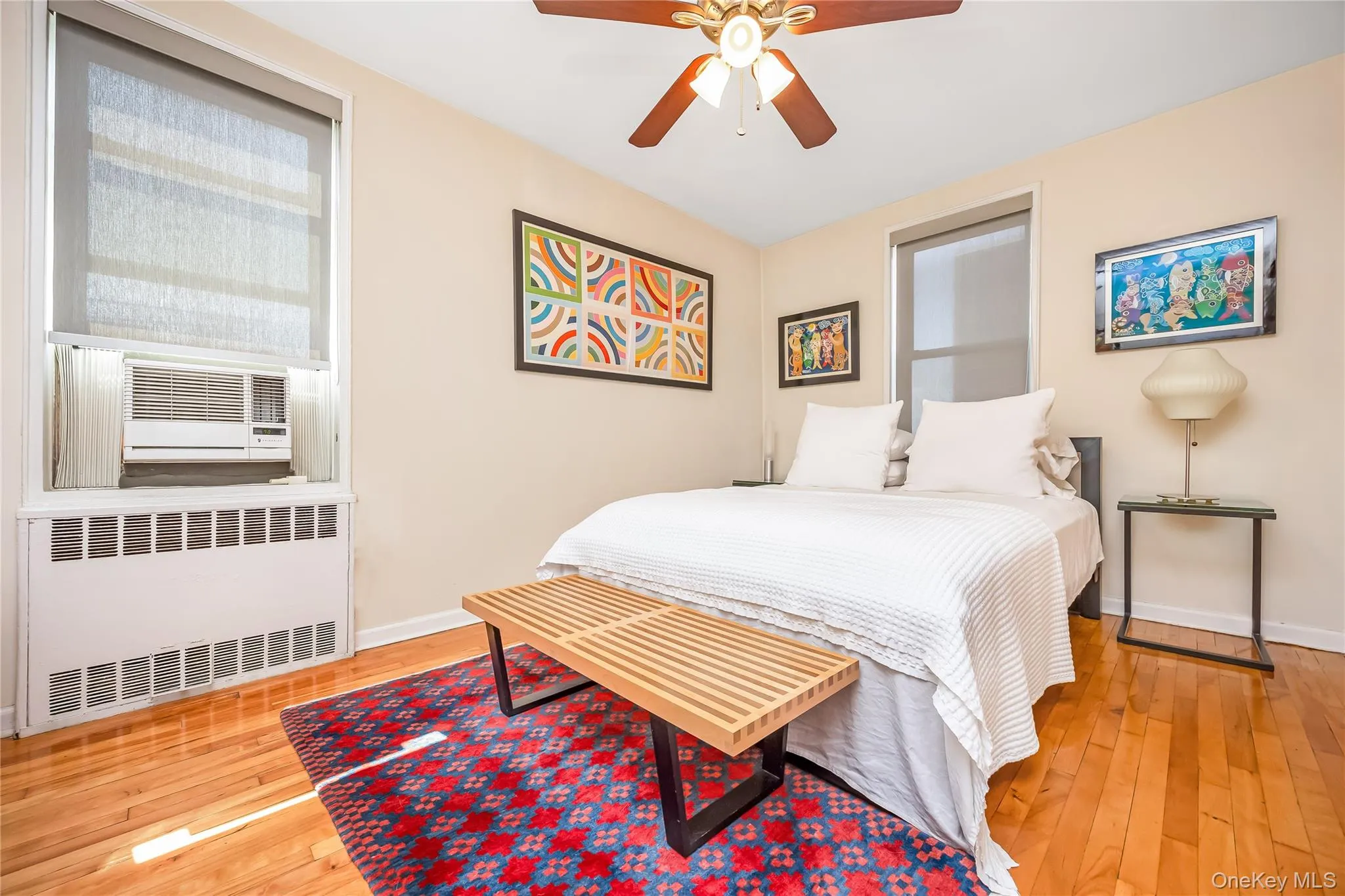 Bright bedroom featuring wood-finish flooring, a ceiling fan with integrated lighting, and a window with a roller shade Bright bedroom featuring wood-finish flooring, a ceiling fan with integrated lighting, and a window with a roller shade