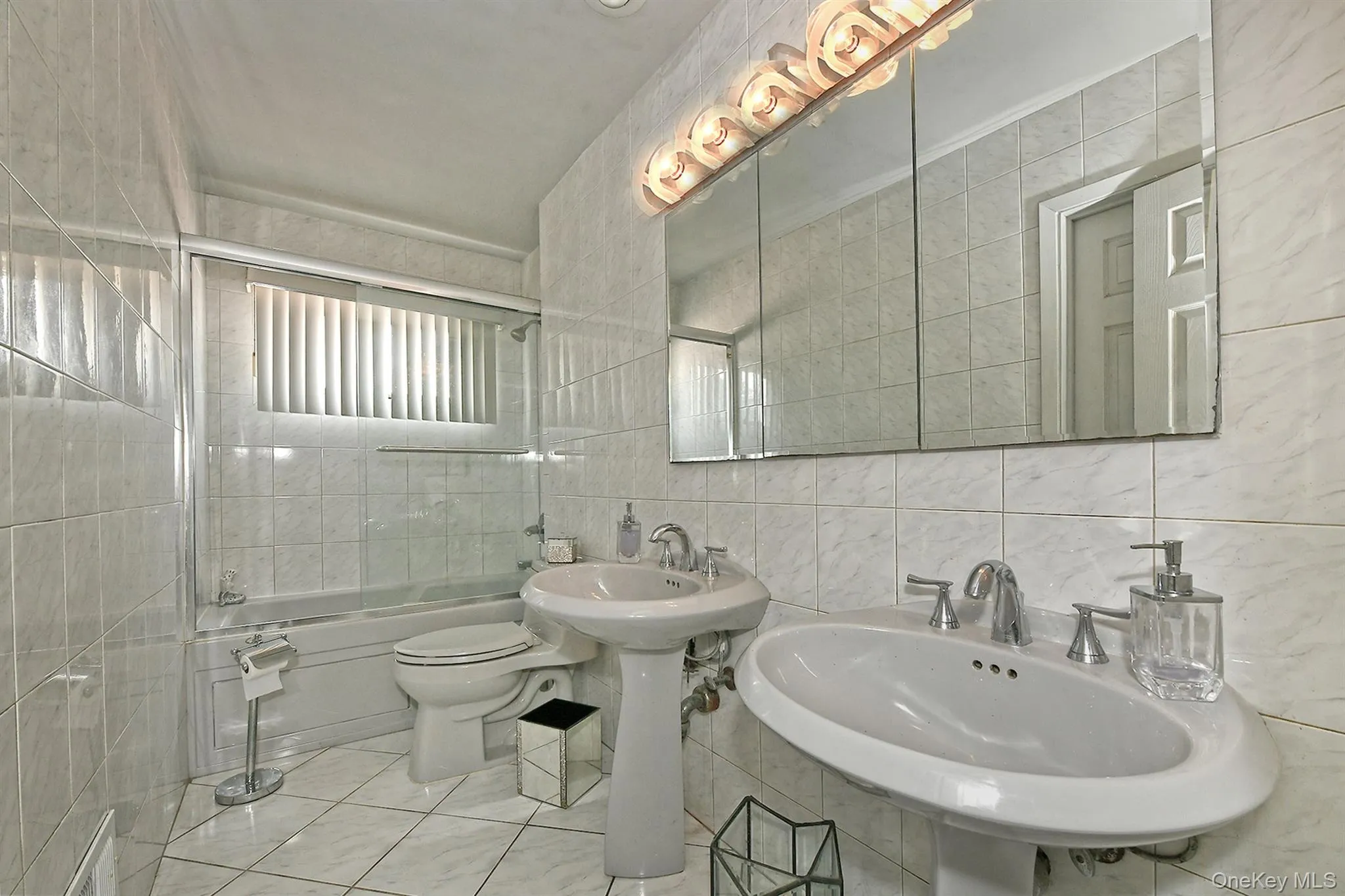 Bathroom featuring dual pedestal sinks, a tiled shower/tub combination, a window with horizontal blinds, a large mirrored medicine cabinet, and overhead vanity lighting Bathroom featuring dual pedestal sinks, a tiled shower/tub combination, a window with horizontal blinds, a large mirrored medicine cabinet, and overhead vanity lighting
