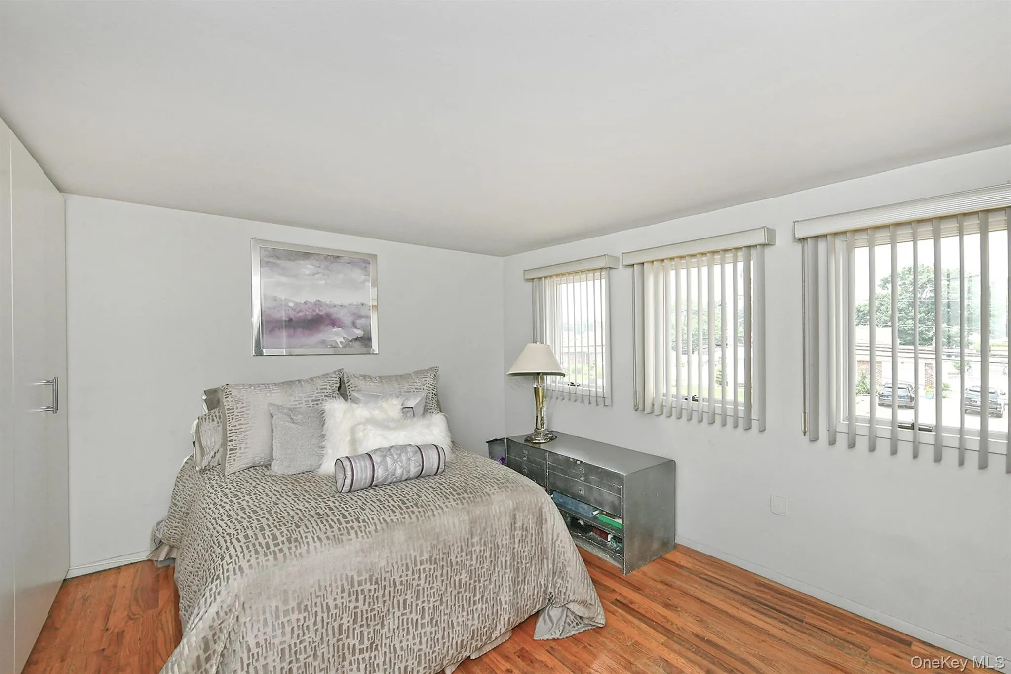 Bright room featuring three windows with vertical blinds, light walls, and rich wood-finish flooring Bright room featuring three windows with vertical blinds, light walls, and rich wood-finish flooring
