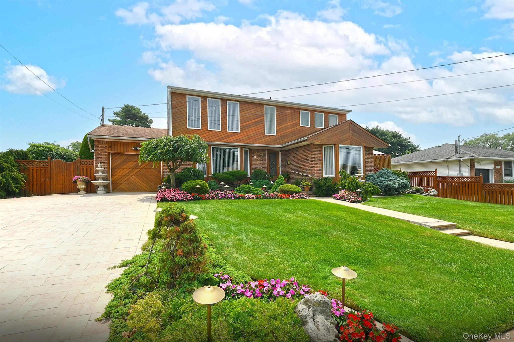 Brick and wood-sided residence featuring a paver driveway, an attached garage with a wood-finish door, a manicured lawn, and extensive landscaping with decorative plantings Brick and wood-sided residence featuring a paver driveway, an attached garage with a wood-finish door, a manicured lawn, and extensive landscaping with decorative plantings