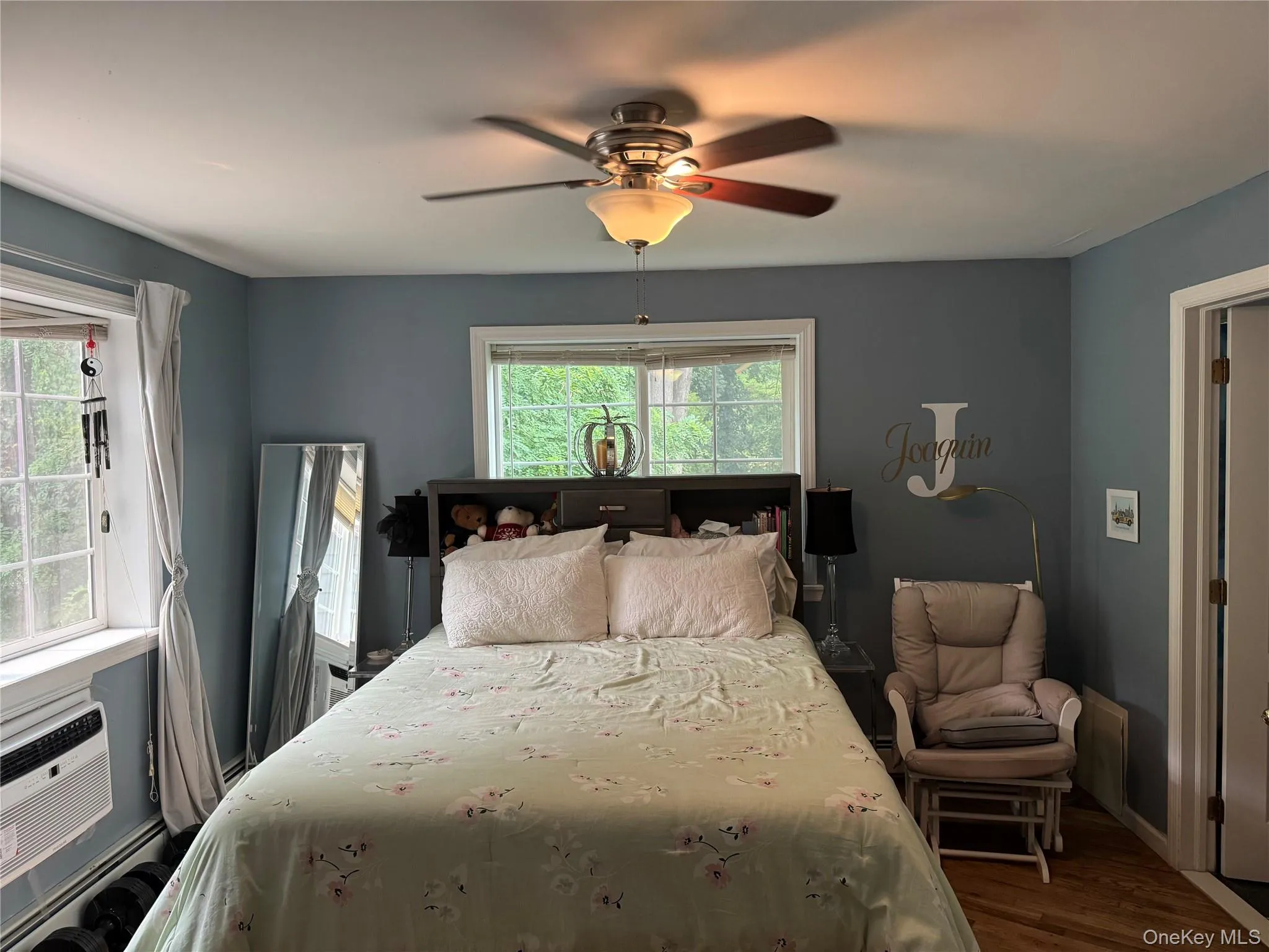 Bedroom featuring two large windows, wood-finish flooring, a ceiling fan with integrated lighting, a built-in headboard storage unit, and a white trim package Bedroom featuring two large windows, wood-finish flooring, a ceiling fan with integrated lighting, a built-in headboard storage unit, and a white trim package