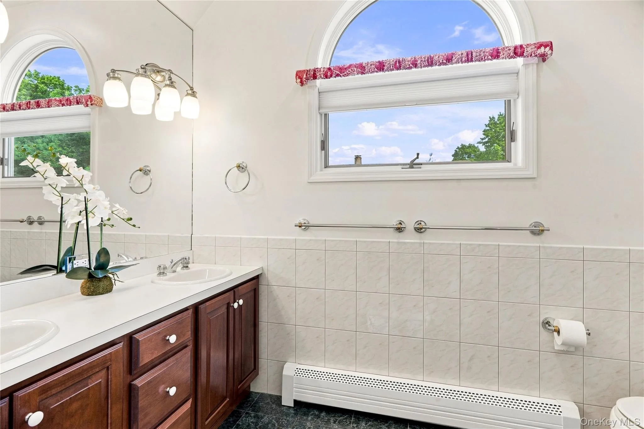 Dual vanity with wood-finish cabinetry, a large wall-mounted mirror, and an arched window Dual vanity with wood-finish cabinetry, a large wall-mounted mirror, and an arched window