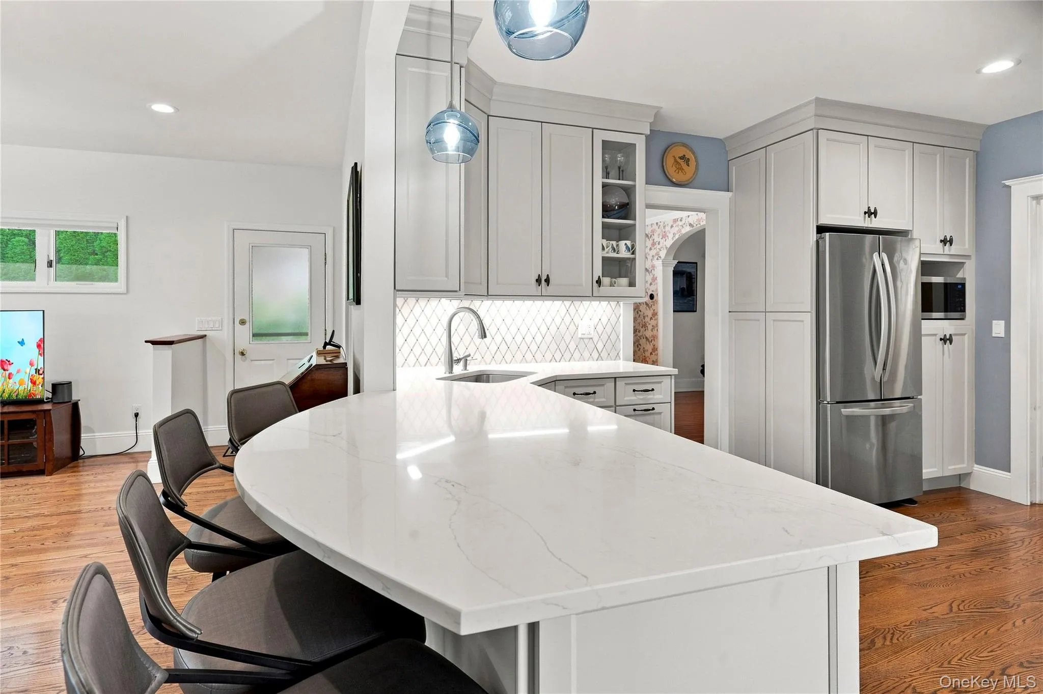Curved kitchen island with a white countertop, light gray cabinetry, stainless steel appliances, pendant lighting, and wood-finish flooring Curved kitchen island with a white countertop, light gray cabinetry, stainless steel appliances, pendant lighting, and wood-finish flooring