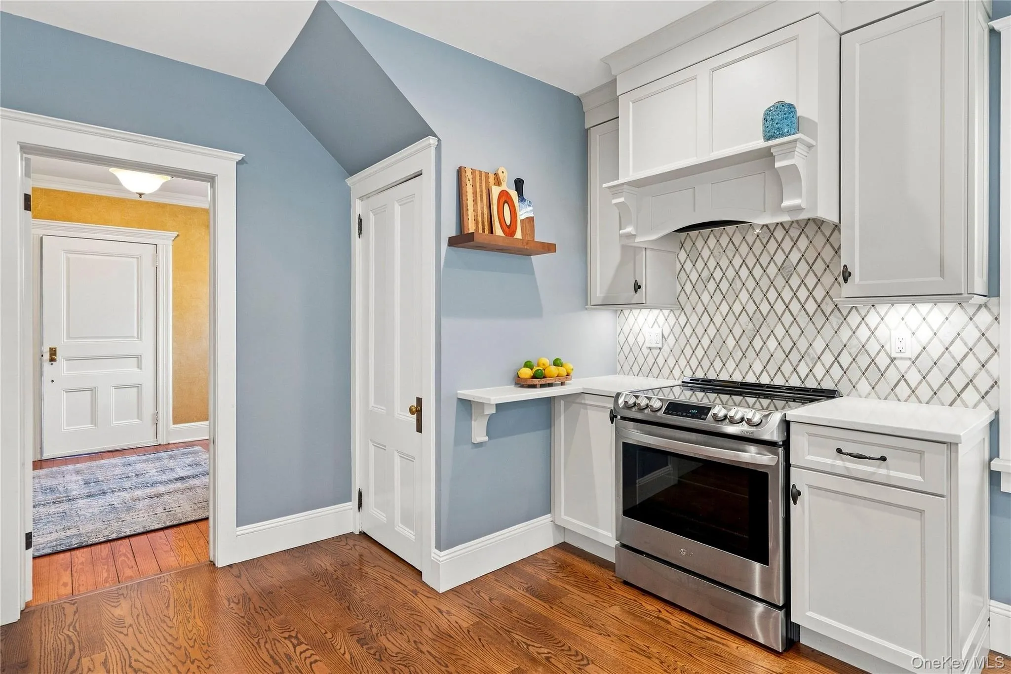 Hardwood flooring, white shaker cabinetry, stainless steel range, patterned marble backsplash, and under-cabinet lighting Hardwood flooring, white shaker cabinetry, stainless steel range, patterned marble backsplash, and under-cabinet lighting