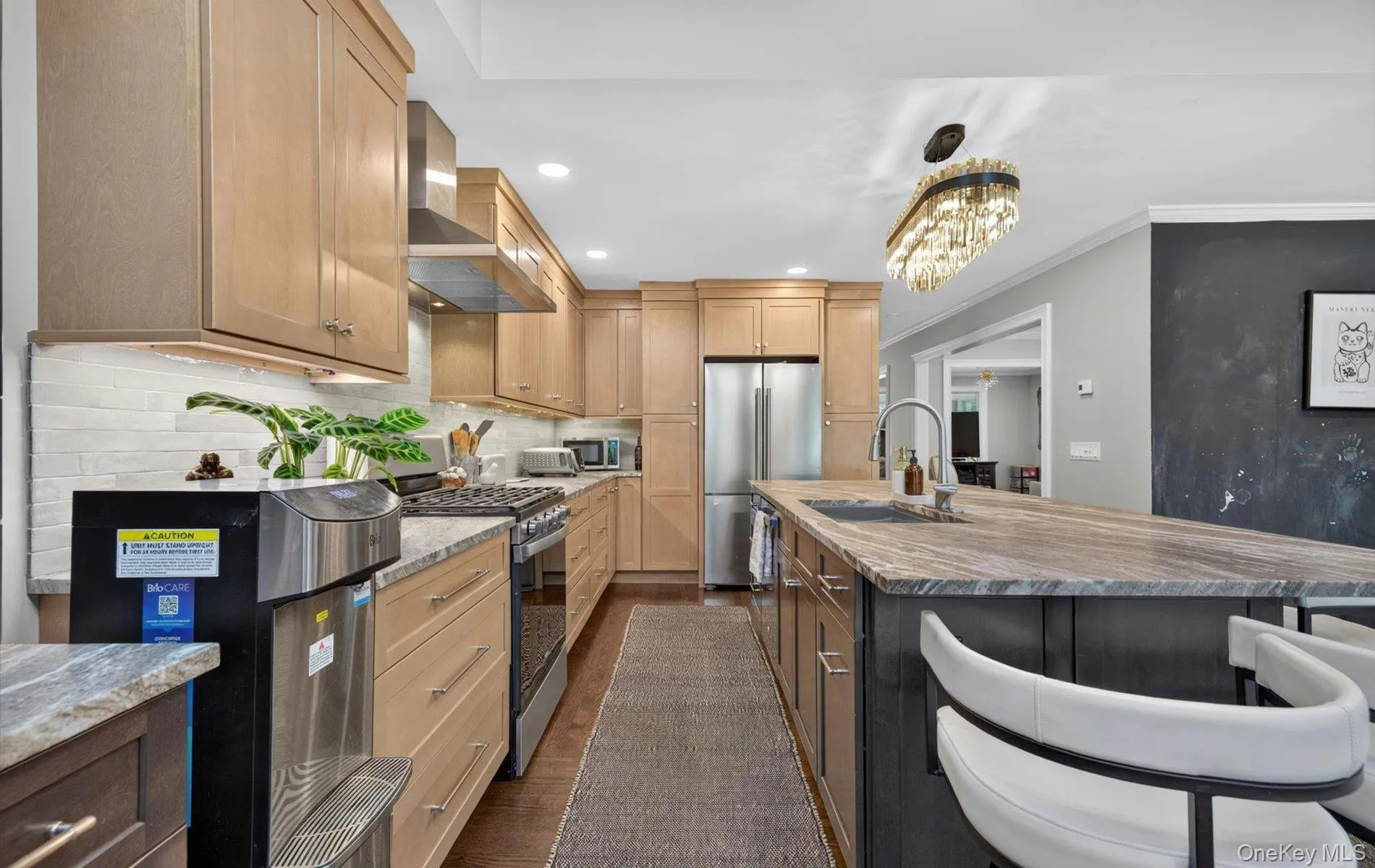 Spacious kitchen featuring light wood cabinetry, stainless steel appliances, a gas range with an overhead vent hood, a large island with a built-in sink, and a unique chandelier light fixture Spacious kitchen featuring light wood cabinetry, stainless steel appliances, a gas range with an overhead vent hood, a large island with a built-in sink, and a unique chandelier light fixture