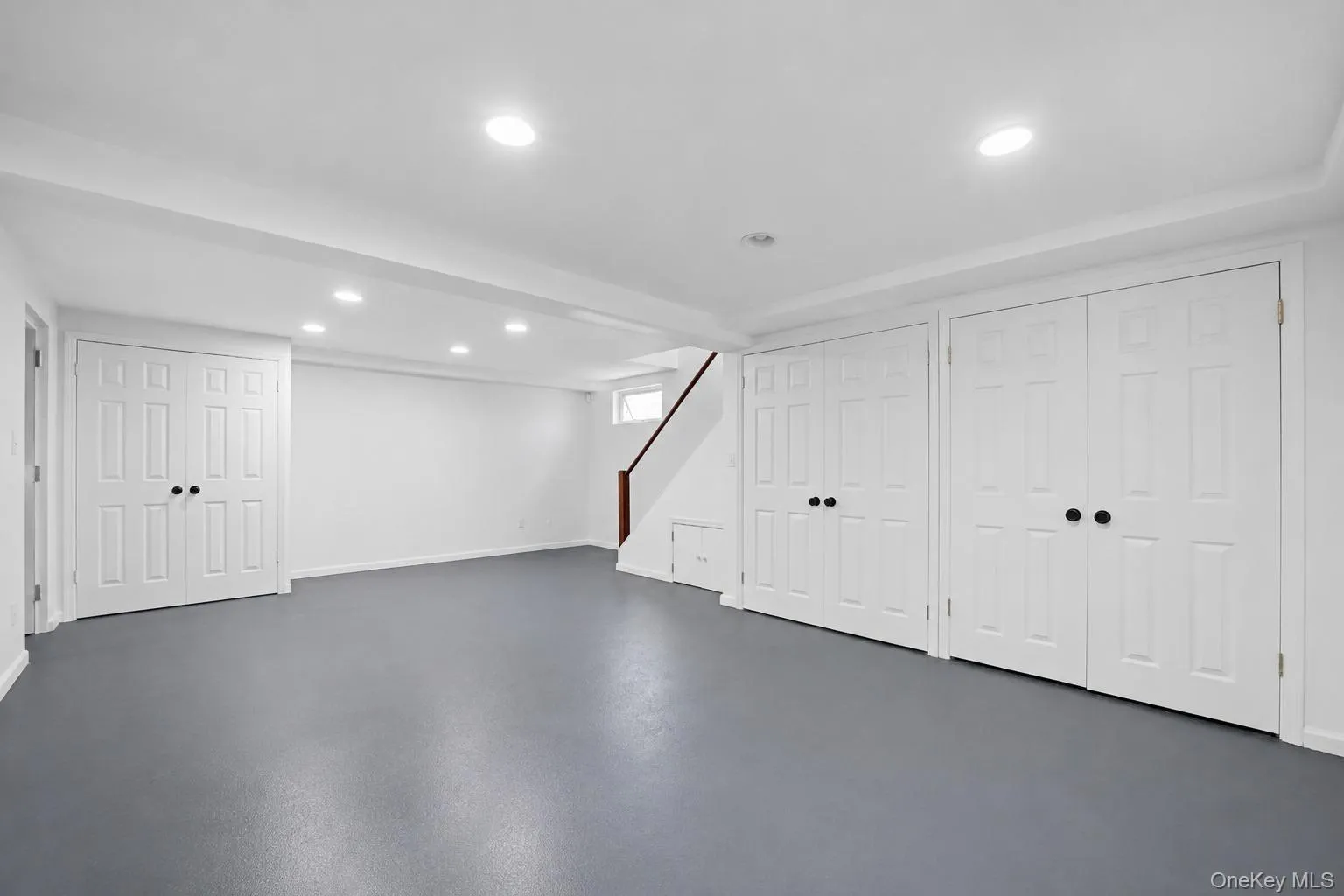 Spacious basement recreation room featuring gray flooring, recessed lighting, multiple paneled closet doors with black hardware, and a staircase with a wood handrail Spacious basement recreation room featuring gray flooring, recessed lighting, multiple paneled closet doors with black hardware, and a staircase with a wood handrail