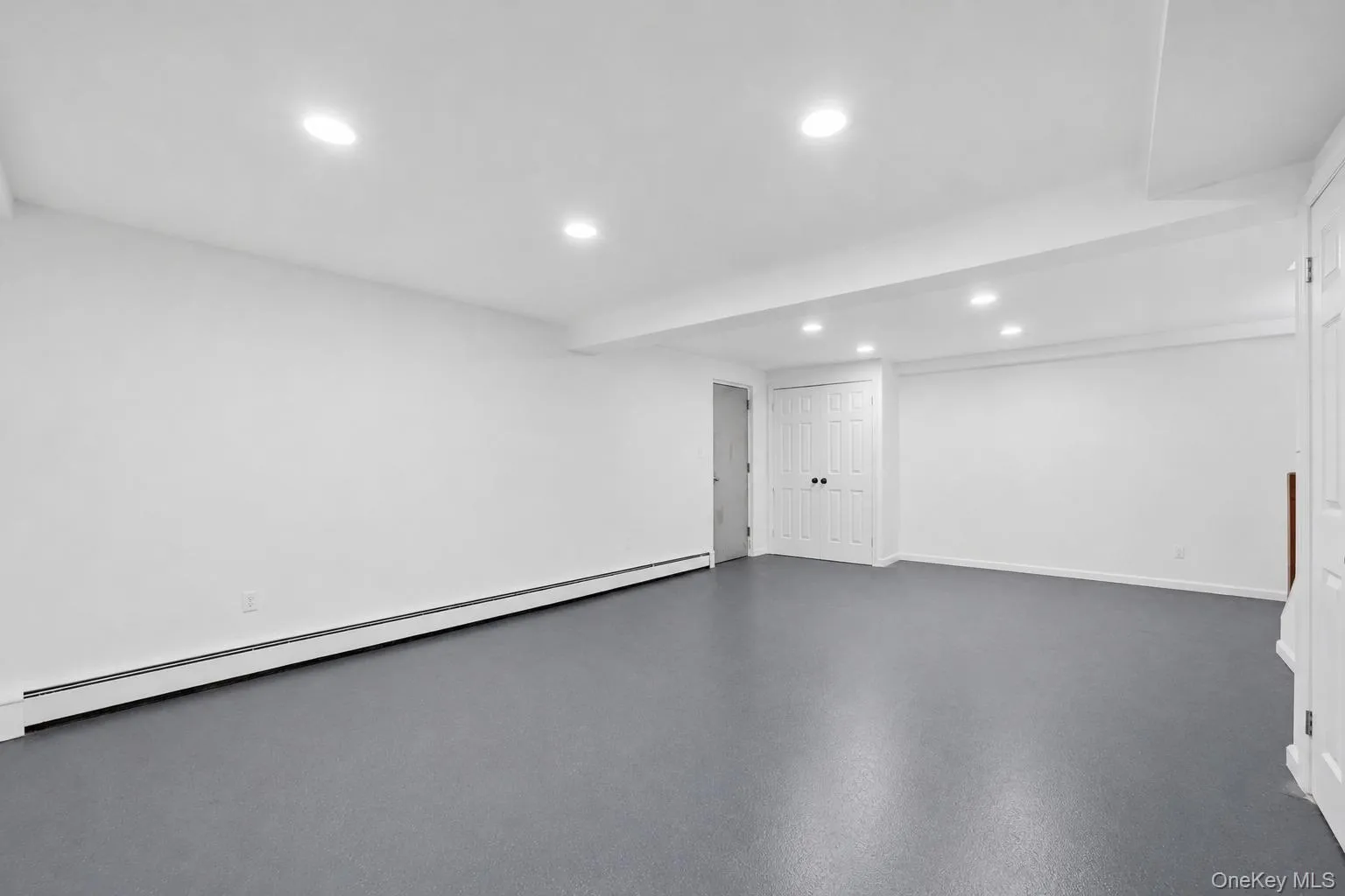 Spacious basement recreation room featuring gray flooring, recessed lighting, multiple paneled closet doors with black hardware, and a staircase with a wood handrail Spacious basement recreation room featuring gray flooring, recessed lighting, multiple paneled closet doors with black hardware, and a staircase with a wood handrail