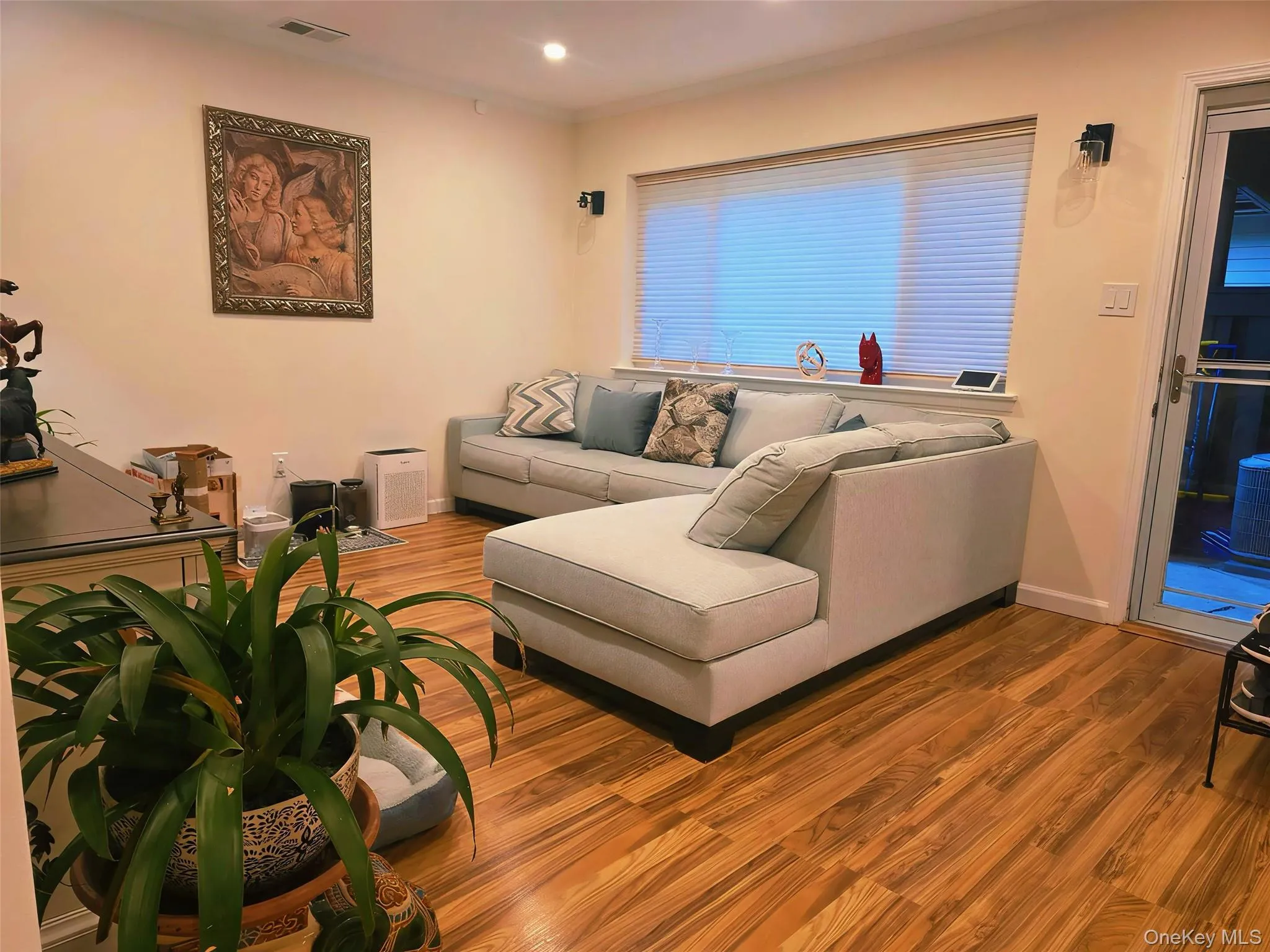 Spacious living area featuring wood-finish flooring, a large window with horizontal blinds, and a glass-paneled exterior door Spacious living area featuring wood-finish flooring, a large window with horizontal blinds, and a glass-paneled exterior door