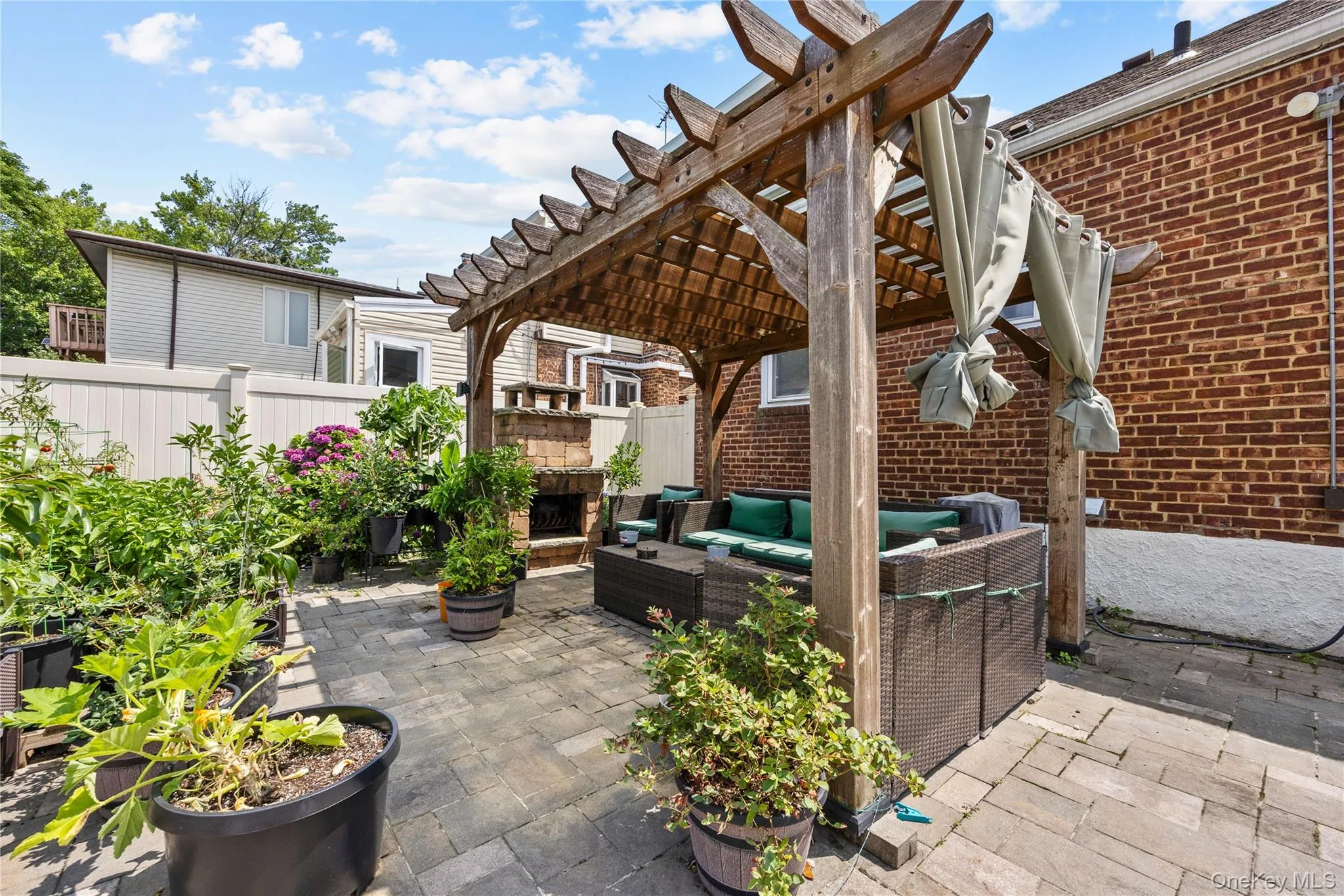 Paved patio featuring a wooden pergola with fabric accents, a built-in stone fireplace, and surrounding garden beds with established greenery Paved patio featuring a wooden pergola with fabric accents, a built-in stone fireplace, and surrounding garden beds with established greenery