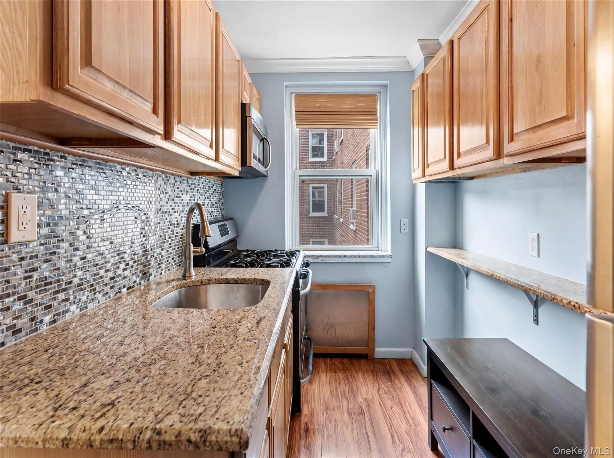 Compact kitchen featuring wood-finish cabinetry, granite-style countertops, a mosaic tile backsplash, and a window providing natural light Compact kitchen featuring wood-finish cabinetry, granite-style countertops, a mosaic tile backsplash, and a window providing natural light