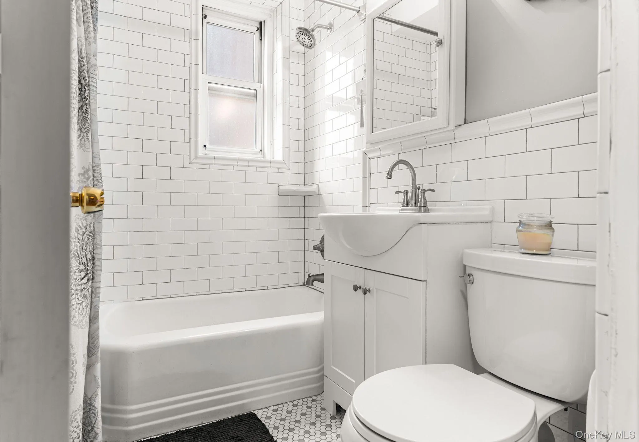 Bathroom featuring white subway tile walls, a built-in bathtub with shower, a vanity sink with cabinet storage, a white toilet, and mosaic tile flooring Bathroom featuring white subway tile walls, a built-in bathtub with shower, a vanity sink with cabinet storage, a white toilet, and mosaic tile flooring