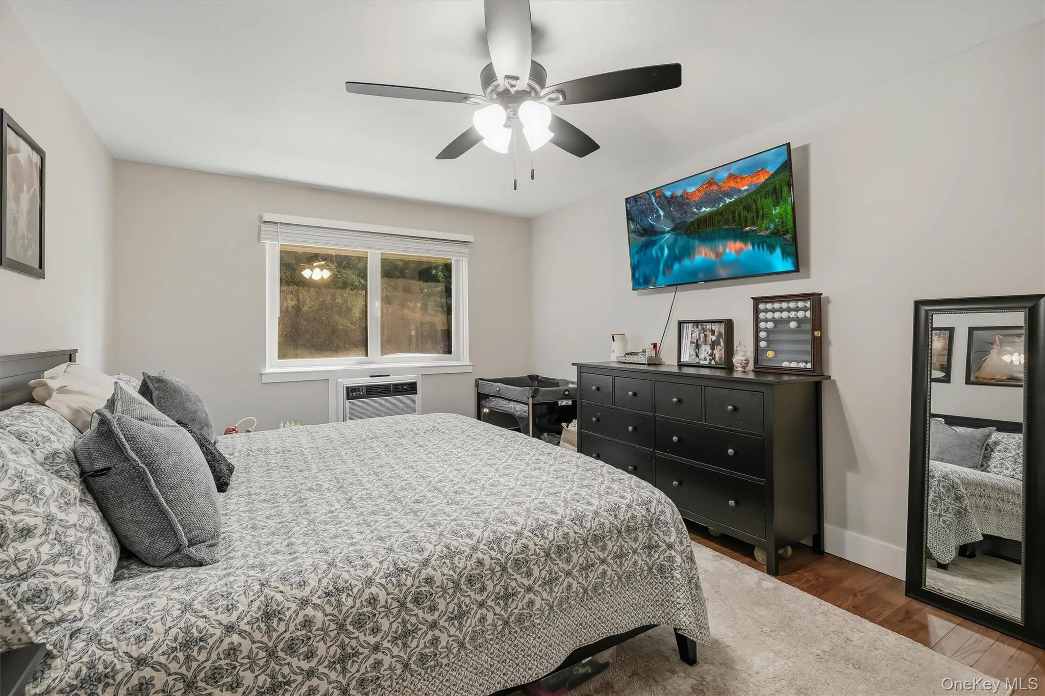 Spacious bedroom featuring wood-finish flooring, a ceiling fan with integrated lighting, and a window with a white frame Spacious bedroom featuring wood-finish flooring, a ceiling fan with integrated lighting, and a window with a white frame