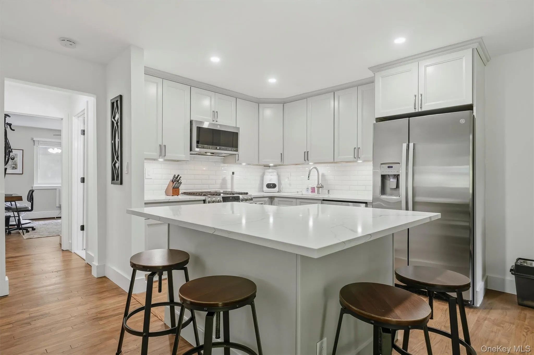 Modern kitchen featuring white shaker-style cabinetry, stainless steel appliances including a built-in microwave and refrigerator, white quartz countertops, a waterfall-edge island with seating for three, stainless steel sink with pull-down faucet, gas cooktop, recessed lighting, and wood-finish flooring throughout Modern kitchen featuring white shaker-style cabinetry, stainless steel appliances including a built-in microwave and refrigerator, white quartz countertops, a waterfall-edge island with seating for three, stainless steel sink with pull-down faucet, gas cooktop, recessed lighting, and wood-finish flooring throughout