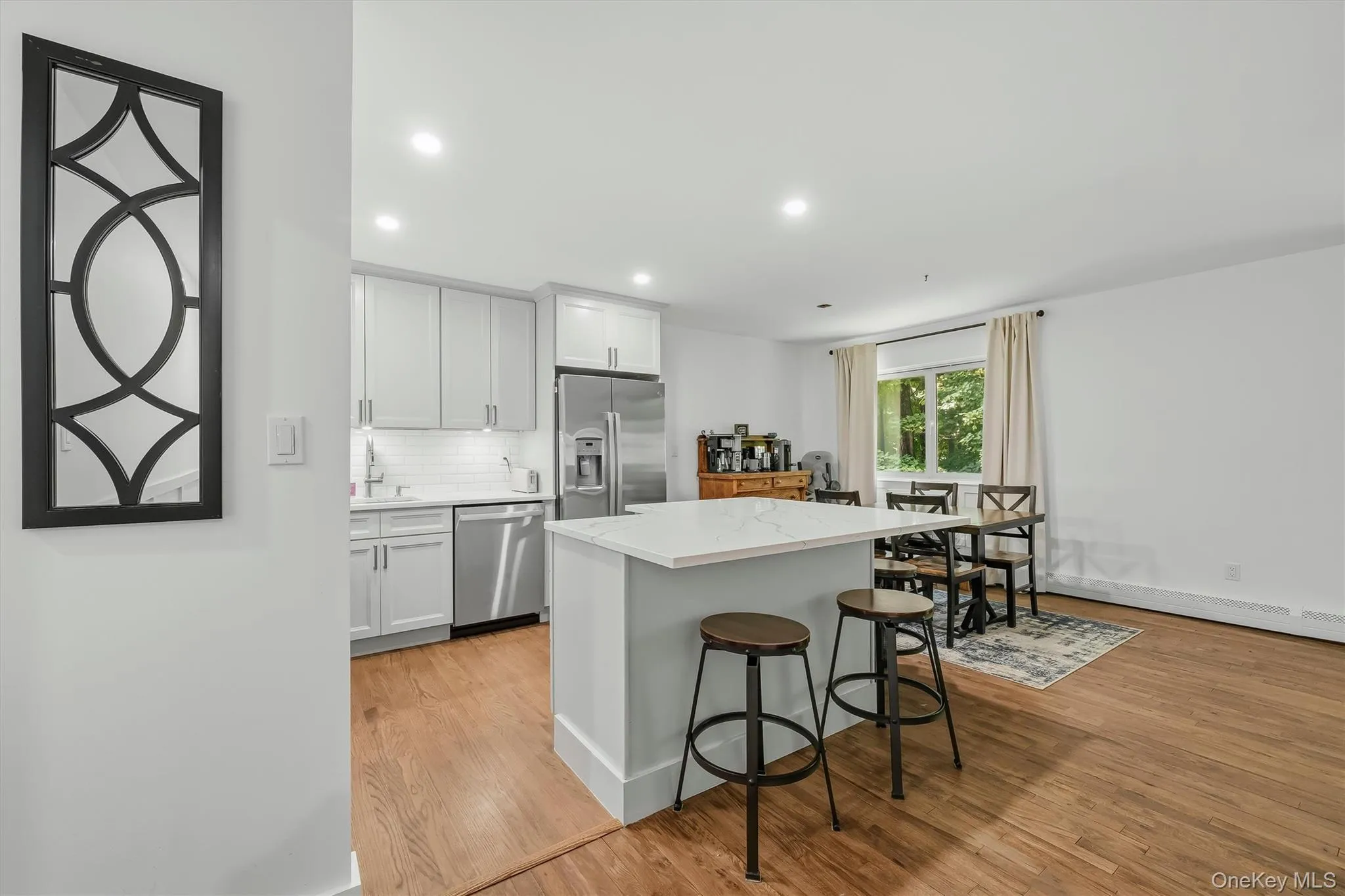 Open-concept kitchen and dining area featuring a white cabinetry kitchen with stainless steel appliances, a white quartz island with seating for two, recessed ceiling lighting, and wood-finish flooring throughout Open-concept kitchen and dining area featuring a white cabinetry kitchen with stainless steel appliances, a white quartz island with seating for two, recessed ceiling lighting, and wood-finish flooring throughout