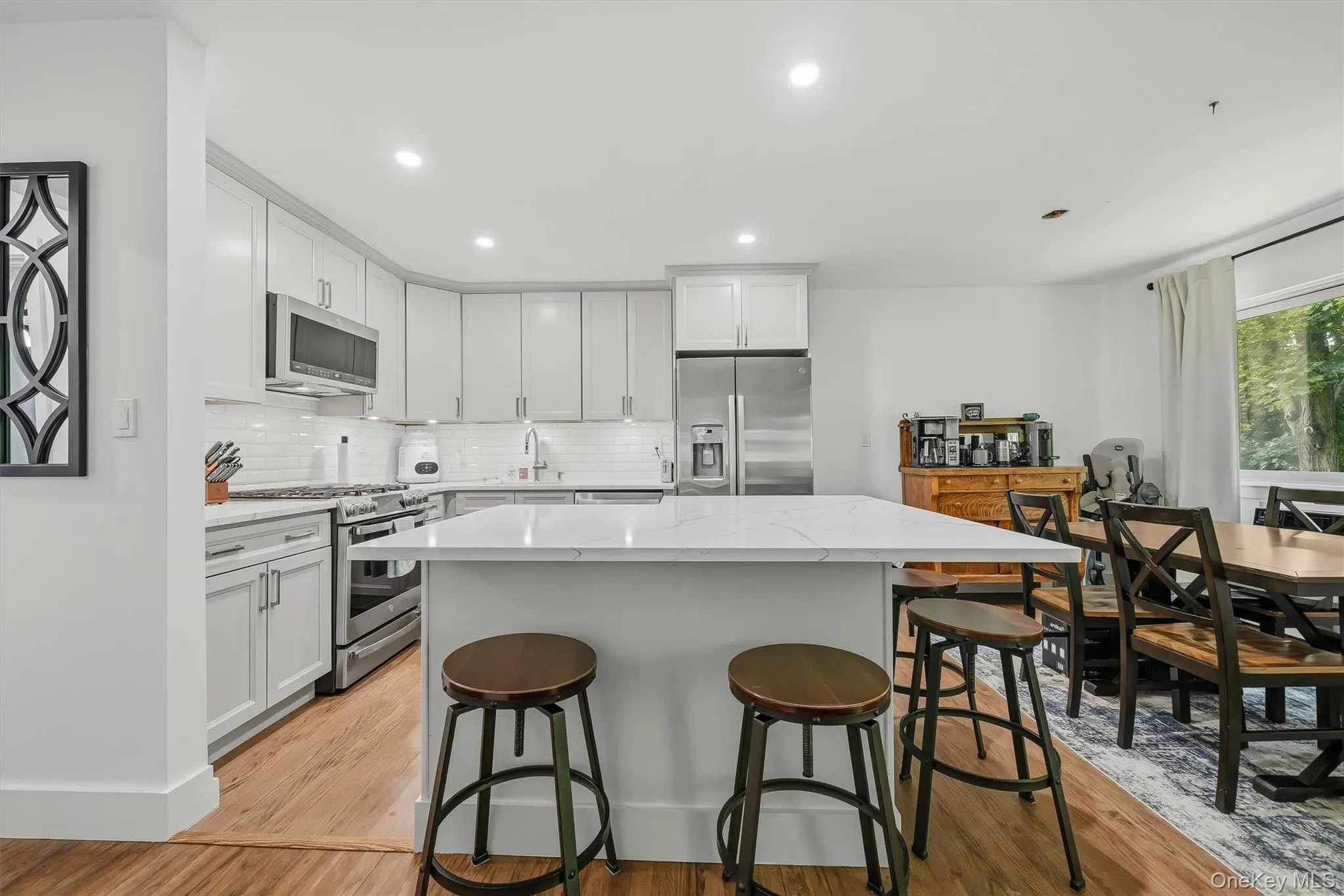 Contemporary kitchen featuring white shaker cabinetry, stainless steel appliances, a white subway tile backsplash, and a large central island with a light-toned countertop Contemporary kitchen featuring white shaker cabinetry, stainless steel appliances, a white subway tile backsplash, and a large central island with a light-toned countertop