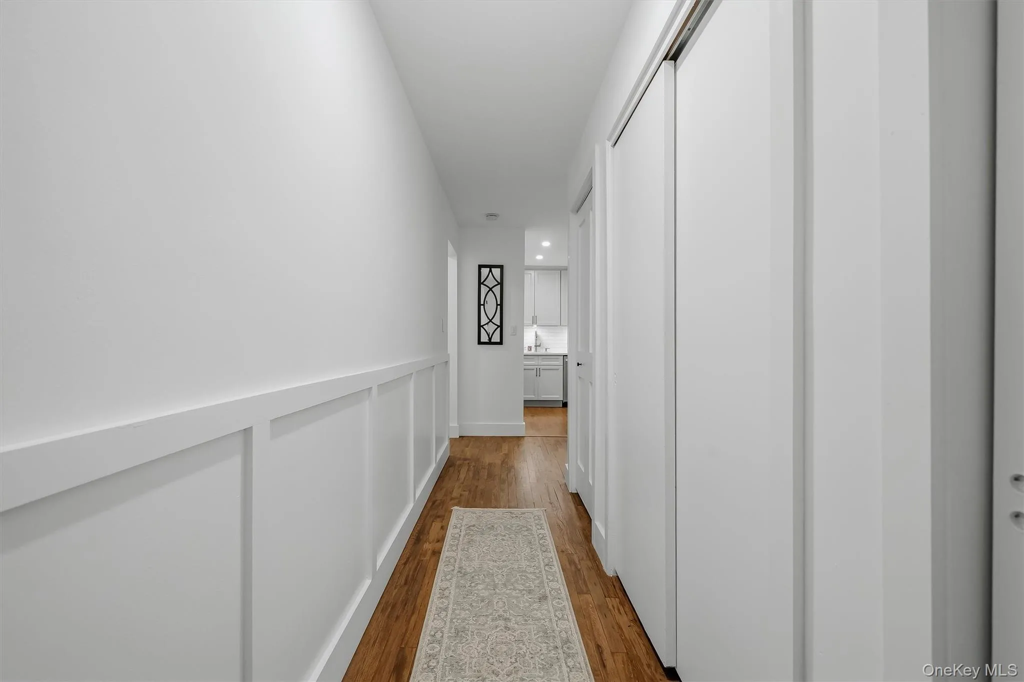 Hallway featuring crisp white wainscoting with a coordinating rail, recessed ceiling lighting, and warm wood-finish flooring throughout Hallway featuring crisp white wainscoting with a coordinating rail, recessed ceiling lighting, and warm wood-finish flooring throughout