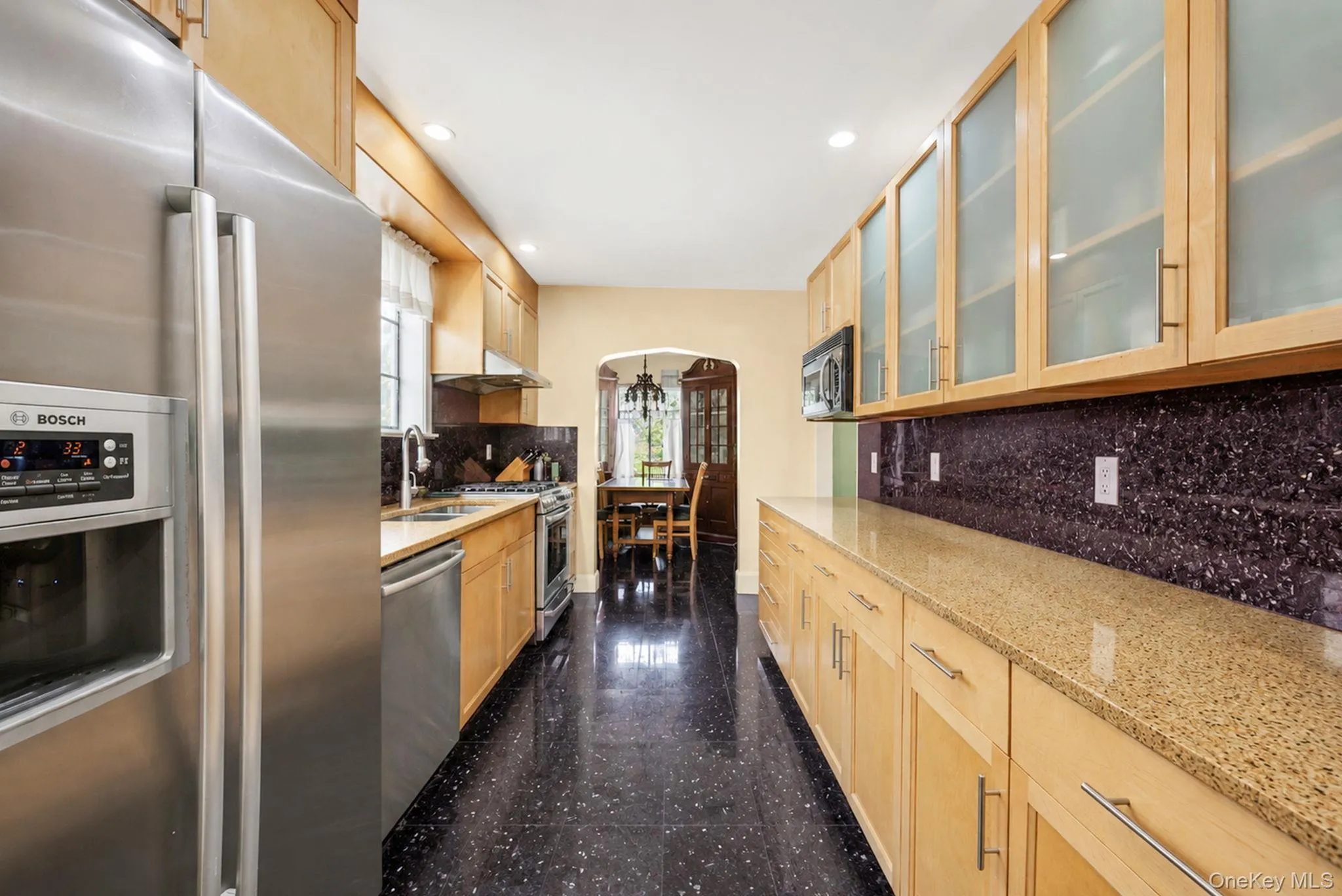 Galley kitchen featuring stainless steel appliances, light wood-finish cabinetry, granite-style countertops, and a dark tile backsplash Galley kitchen featuring stainless steel appliances, light wood-finish cabinetry, granite-style countertops, and a dark tile backsplash