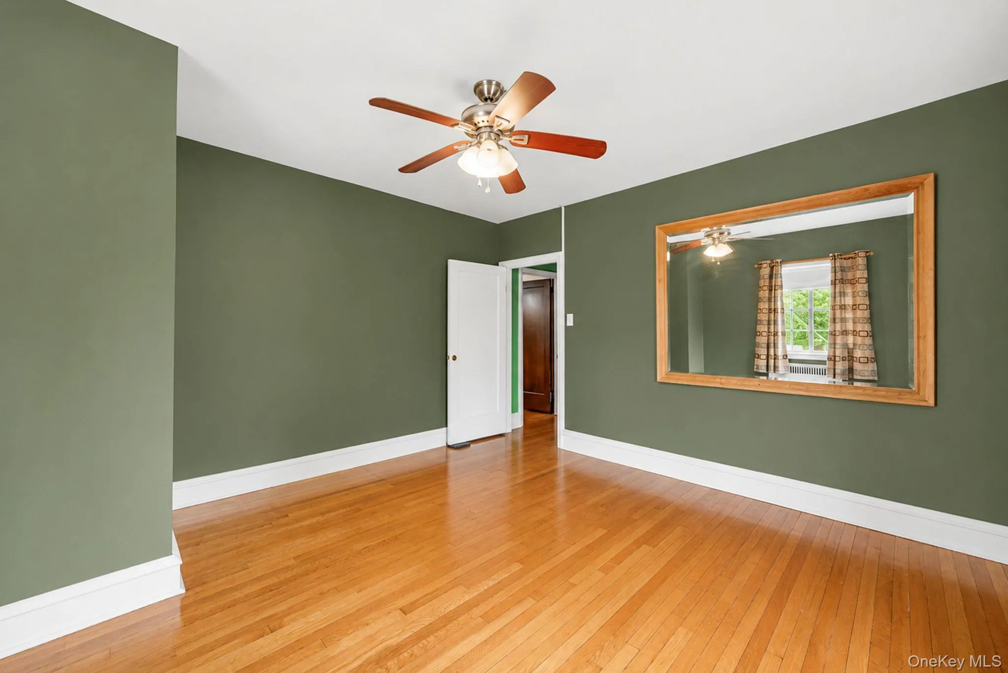 Spacious room featuring wood-finish flooring, a ceiling fan with integrated lighting, and a large wall-mounted mirror with a wood frame Spacious room featuring wood-finish flooring, a ceiling fan with integrated lighting, and a large wall-mounted mirror with a wood frame