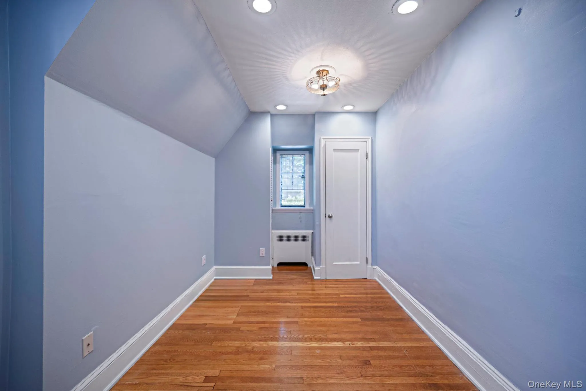 Hardwood flooring and recessed lighting illuminate this interior space, featuring a window with a radiator below Hardwood flooring and recessed lighting illuminate this interior space, featuring a window with a radiator below