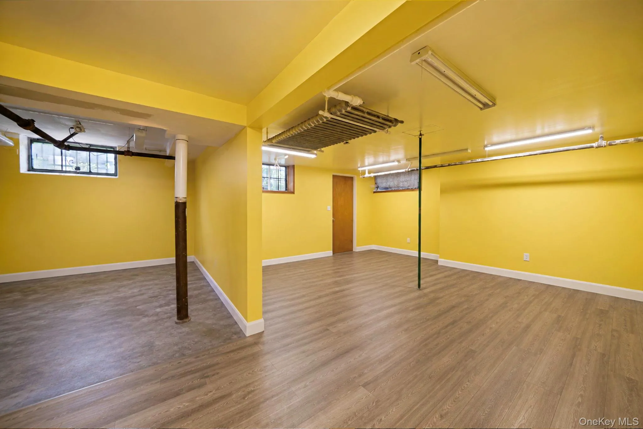 Lower level featuring wood-finish flooring, bright yellow walls, white trim, and multiple fluorescent lighting fixtures Lower level featuring wood-finish flooring, bright yellow walls, white trim, and multiple fluorescent lighting fixtures