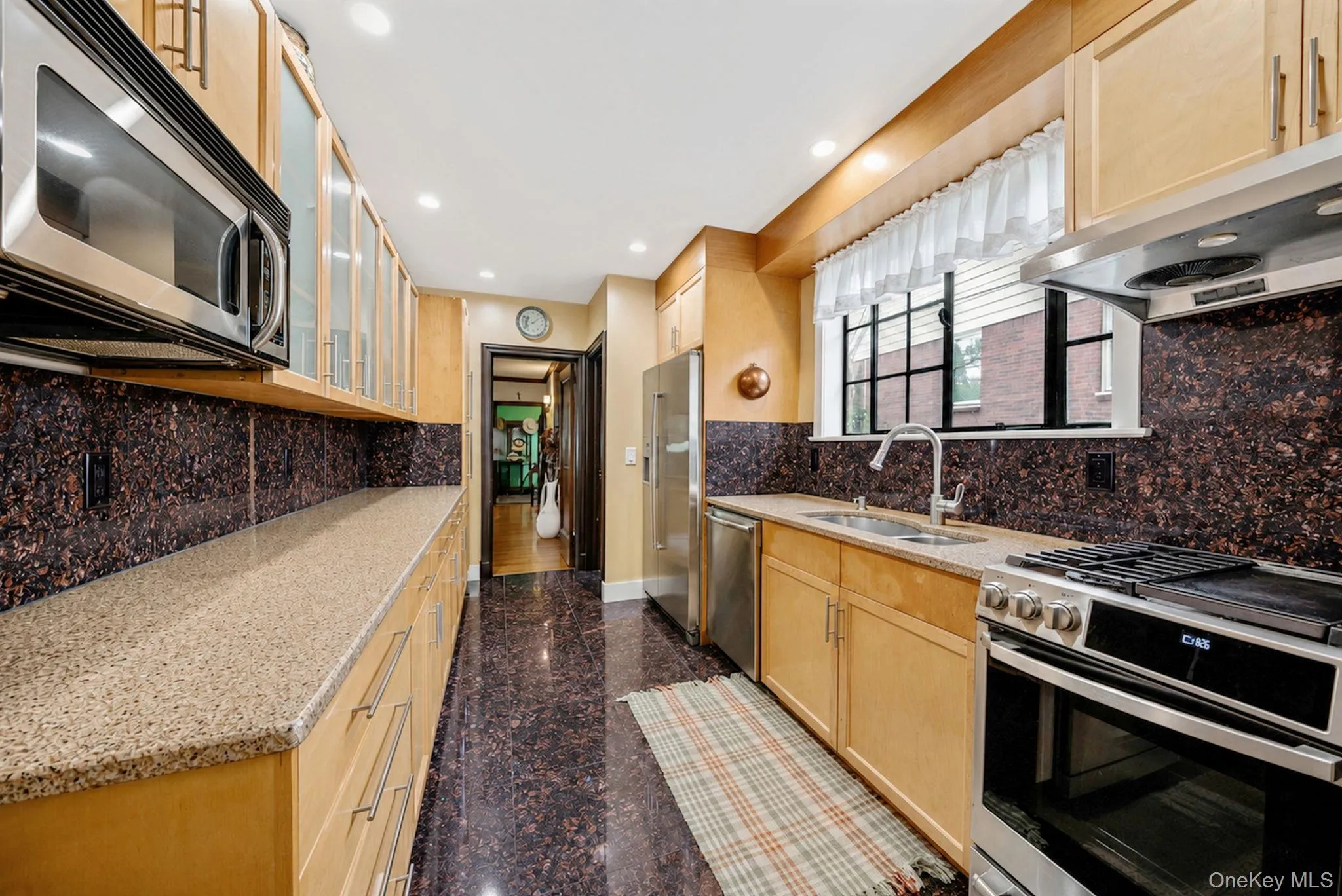 Galley kitchen featuring light wood cabinetry, stone countertops, stainless steel appliances, and a dark stone backsplash Galley kitchen featuring light wood cabinetry, stone countertops, stainless steel appliances, and a dark stone backsplash