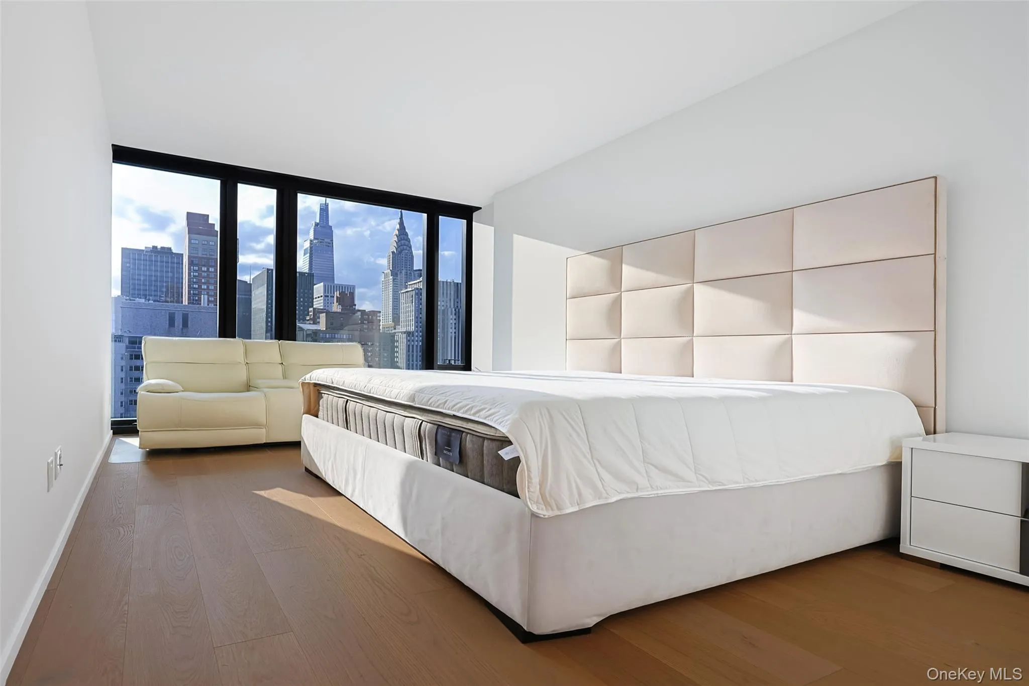 Primary Bedroom with Expansive windows showcasing cityscape views, light wood-finish flooring, white walls, and a paneled upholstered headboard Primary Bedroom with Expansive windows showcasing cityscape views, light wood-finish flooring, white walls, and a paneled upholstered headboard