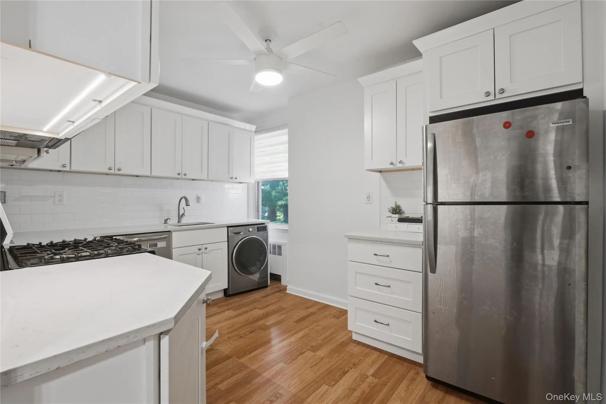 Modern kitchen featuring white shaker cabinetry, stainless steel appliances, white subway tile backsplash, light-toned countertops, and wood-finish flooring Modern kitchen featuring white shaker cabinetry, stainless steel appliances, white subway tile backsplash, light-toned countertops, and wood-finish flooring