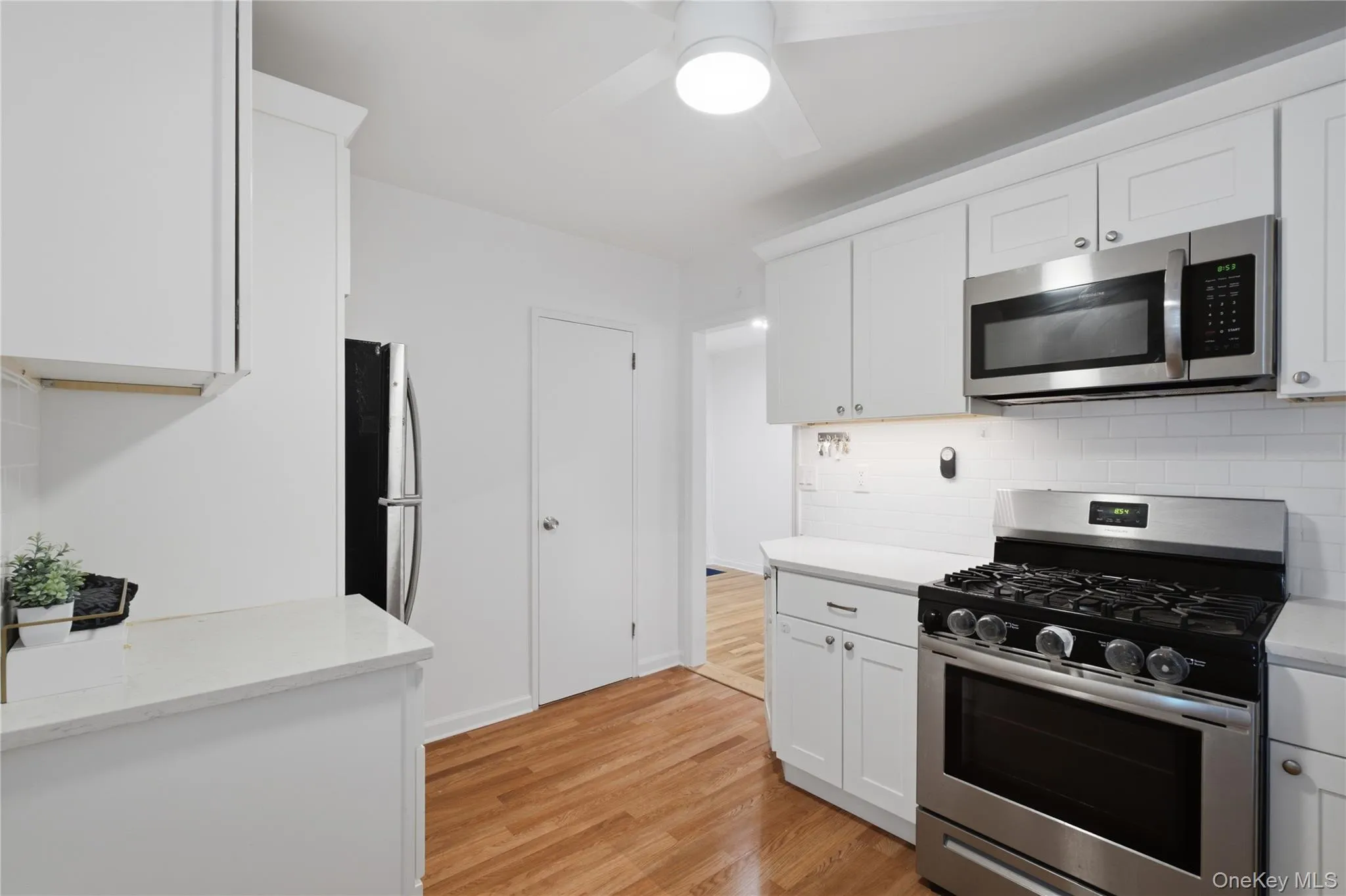 Bright kitchen featuring white shaker cabinetry, stainless steel appliances, a subway tile backsplash, and wood-finish flooring Bright kitchen featuring white shaker cabinetry, stainless steel appliances, a subway tile backsplash, and wood-finish flooring