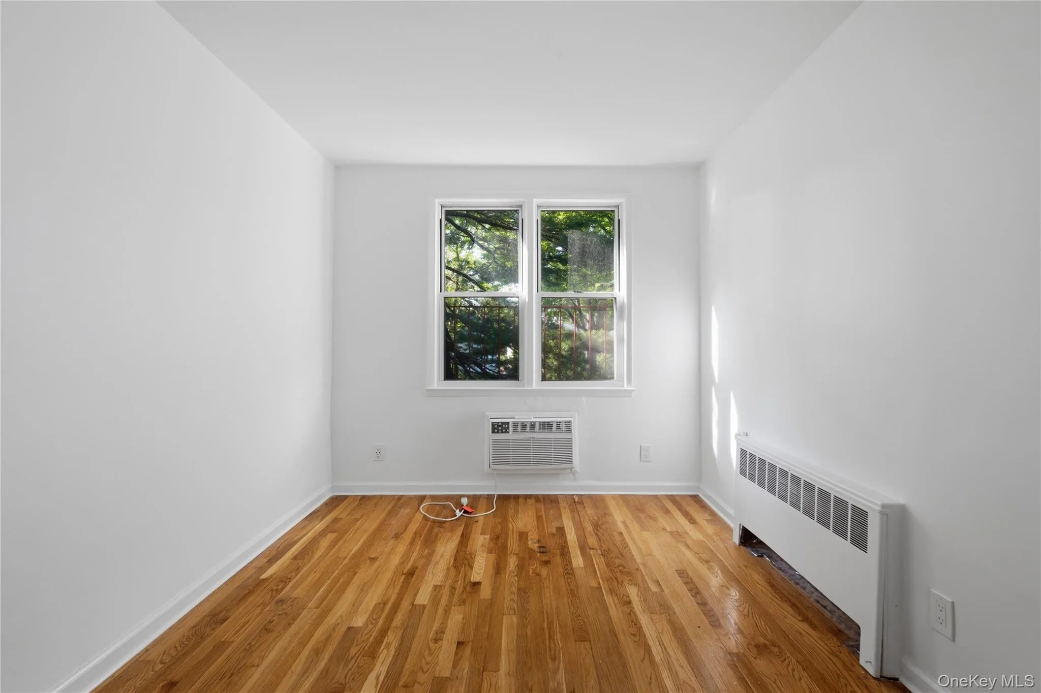 Hardwood flooring in a bright room featuring a window with exterior tree views, a window-mounted air conditioning unit, and a baseboard heating unit Hardwood flooring in a bright room featuring a window with exterior tree views, a window-mounted air conditioning unit, and a baseboard heating unit