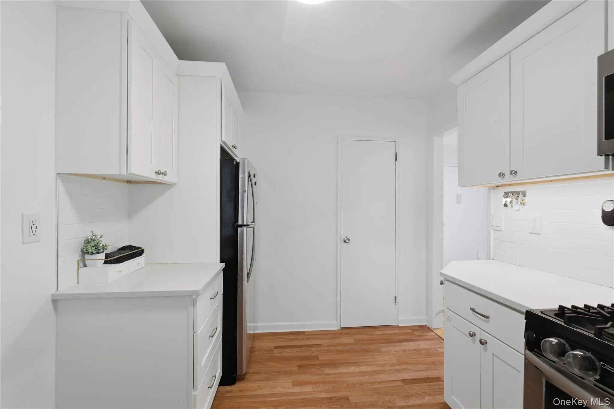 Modern kitchen featuring white shaker cabinetry, light countertops, subway tile backsplash, stainless steel refrigerator, and wood-finish flooring Modern kitchen featuring white shaker cabinetry, light countertops, subway tile backsplash, stainless steel refrigerator, and wood-finish flooring