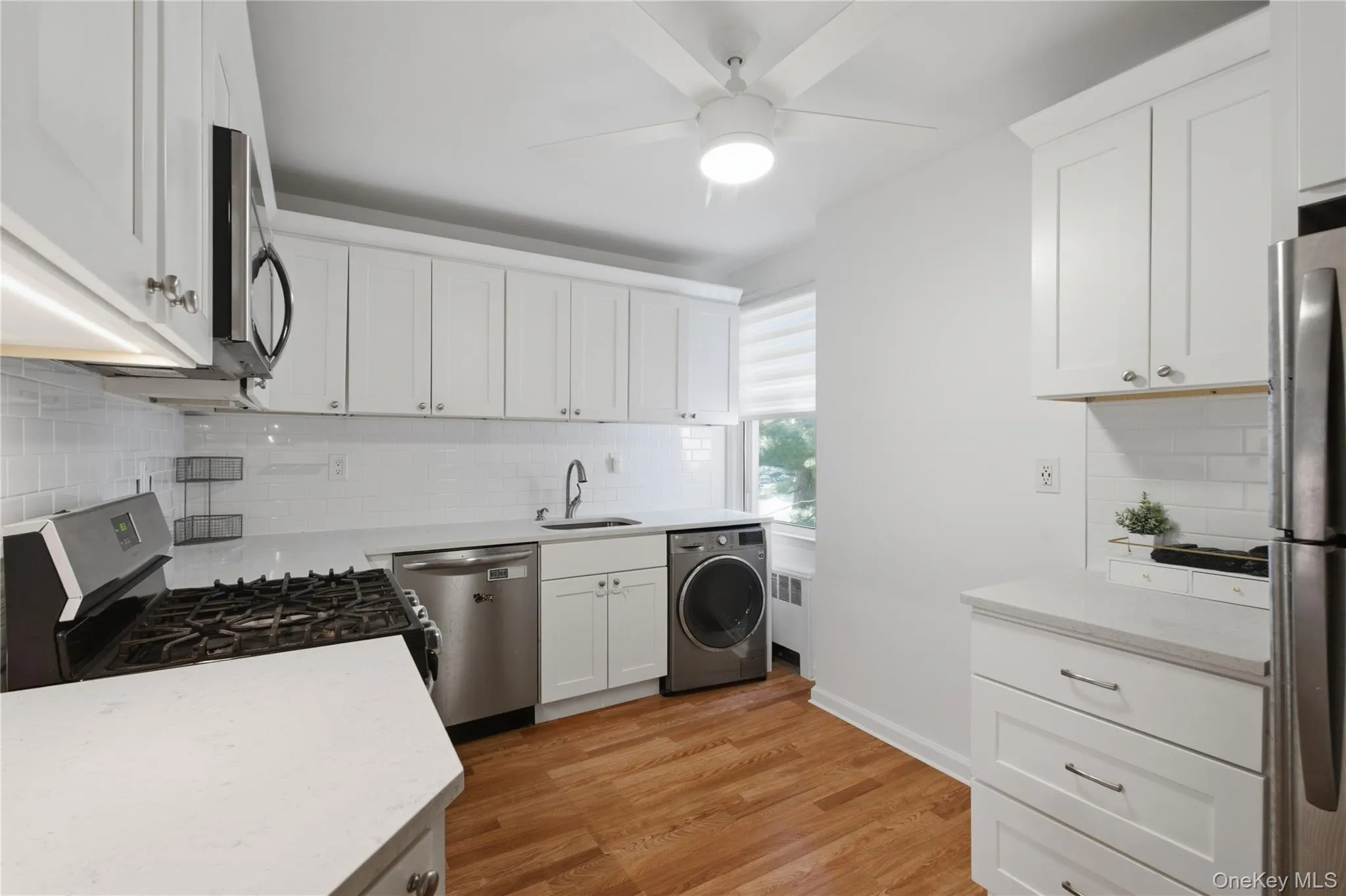 Bright kitchen featuring white shaker cabinetry, stainless steel appliances, white subway tile backsplash, and wood-finish flooring Bright kitchen featuring white shaker cabinetry, stainless steel appliances, white subway tile backsplash, and wood-finish flooring