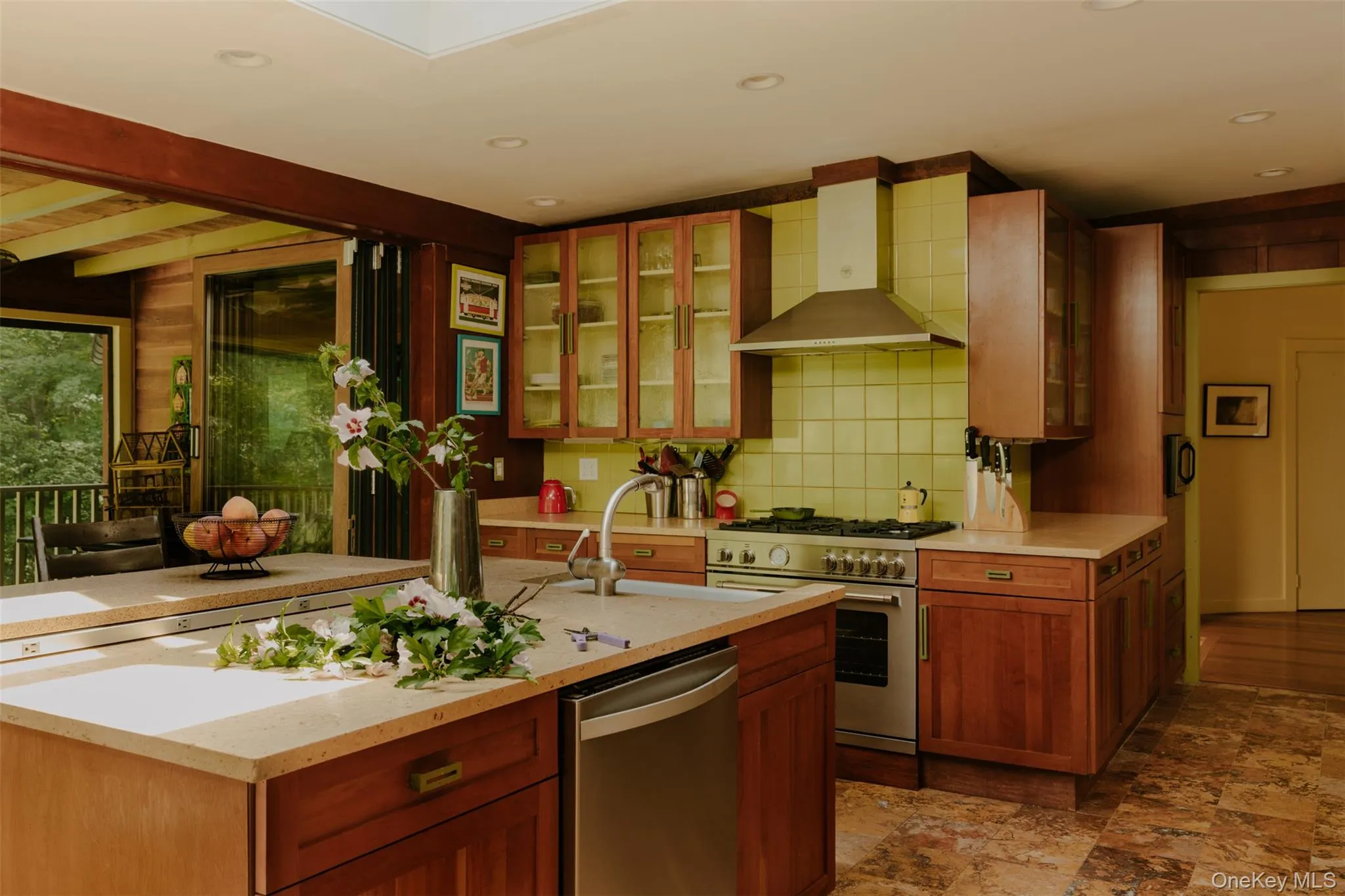Large kitchen featuring an expansive central island with integrated dishwasher, light-toned countertops, rich wood cabinetry, and a stainless steel professional-grade range with a matching vent hood Large kitchen featuring an expansive central island with integrated dishwasher, light-toned countertops, rich wood cabinetry, and a stainless steel professional-grade range with a matching vent hood