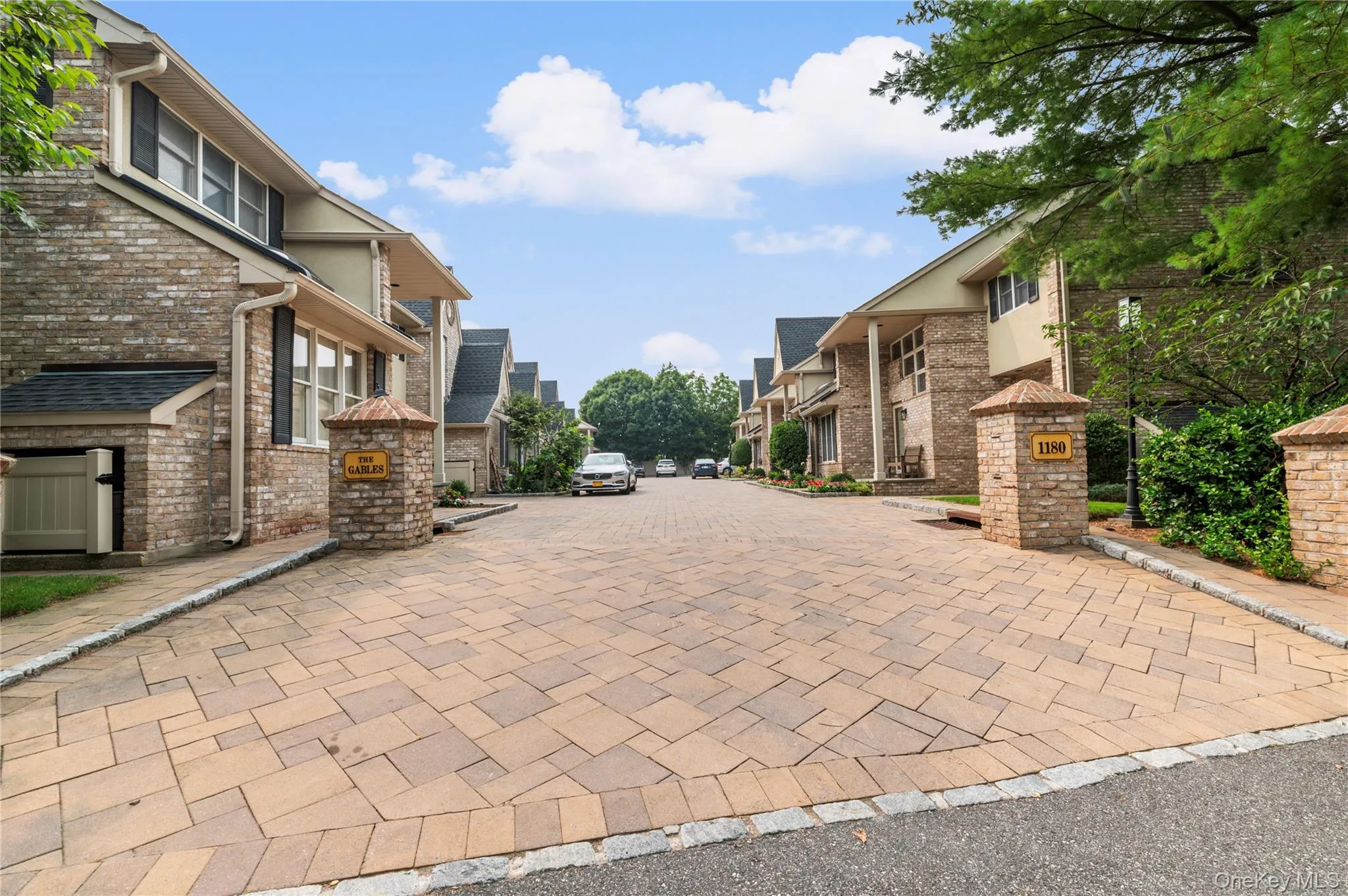 Paved driveway entry featuring stone pillars, brick facades, gable roofs, and mature trees Paved driveway entry featuring stone pillars, brick facades, gable roofs, and mature trees