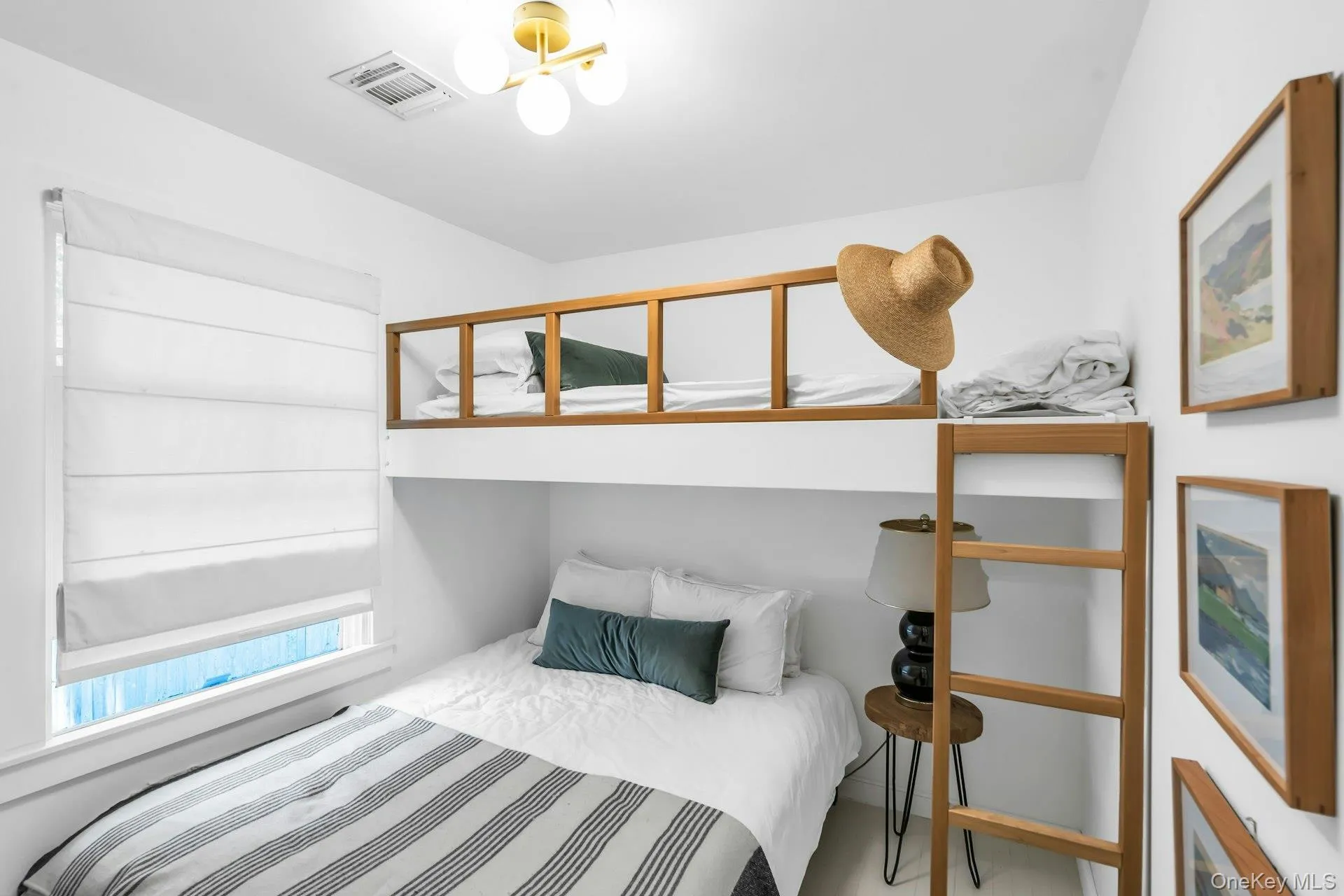 Light-filled room featuring a built-in bunk bed system with natural wood accents, a window with a white Roman shade, a brass ceiling light fixture, and framed wall art Light-filled room featuring a built-in bunk bed system with natural wood accents, a window with a white Roman shade, a brass ceiling light fixture, and framed wall art